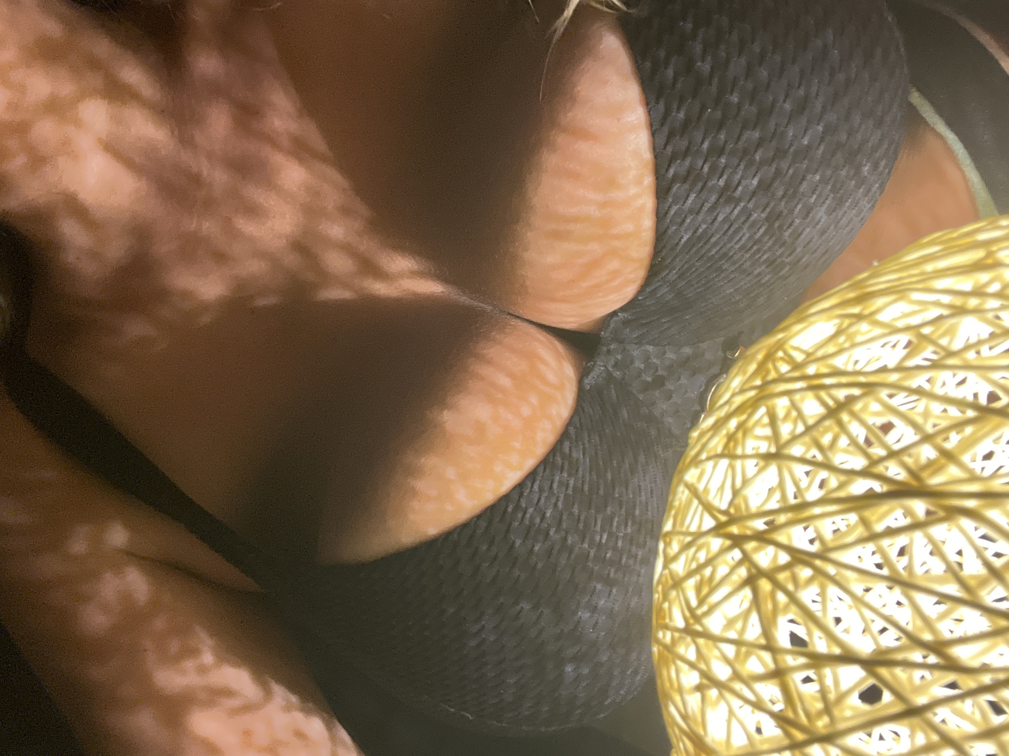 A close-up, overhead shot shows the cleavage of a person wearing a dark, textured bra. The person's skin is visible, with dappled light creating a patterned effect. In the foreground, a large, yellow ball of interwoven string or yarn is illuminated from within. The background is dark and indistinct.