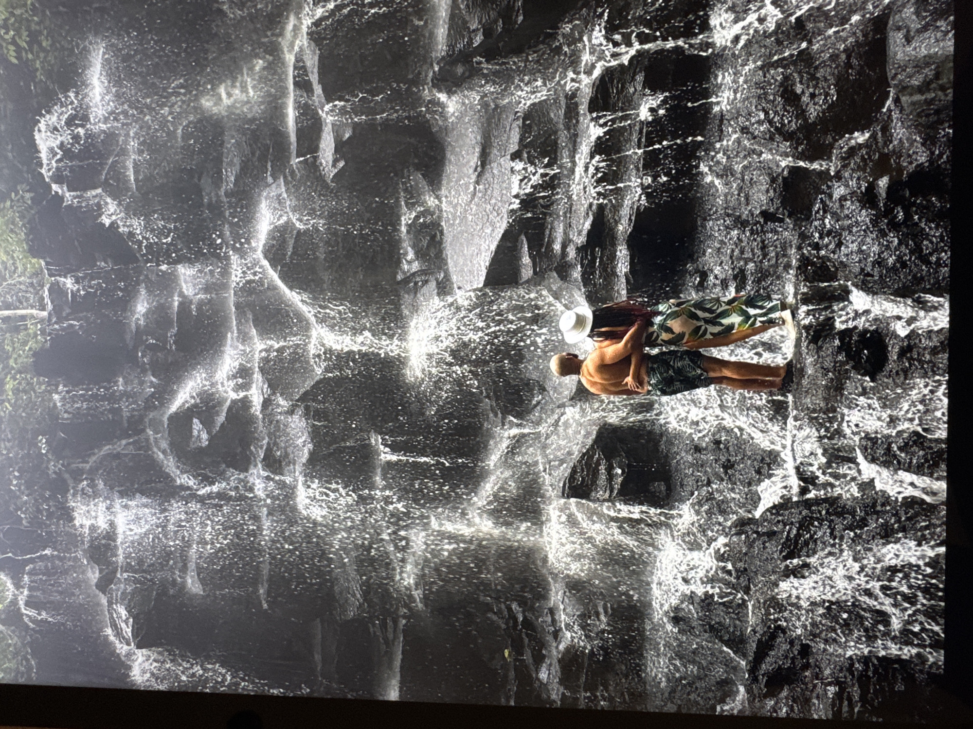 A photograph depicts a waterfall cascading down a dark, rocky cliff. A couple stands on a rocky ledge near the waterfall. The man is shirtless, wearing dark shorts. The woman is wearing a white hat and a floral-patterned dress or cover-up. The location is Unknown, Unknown.