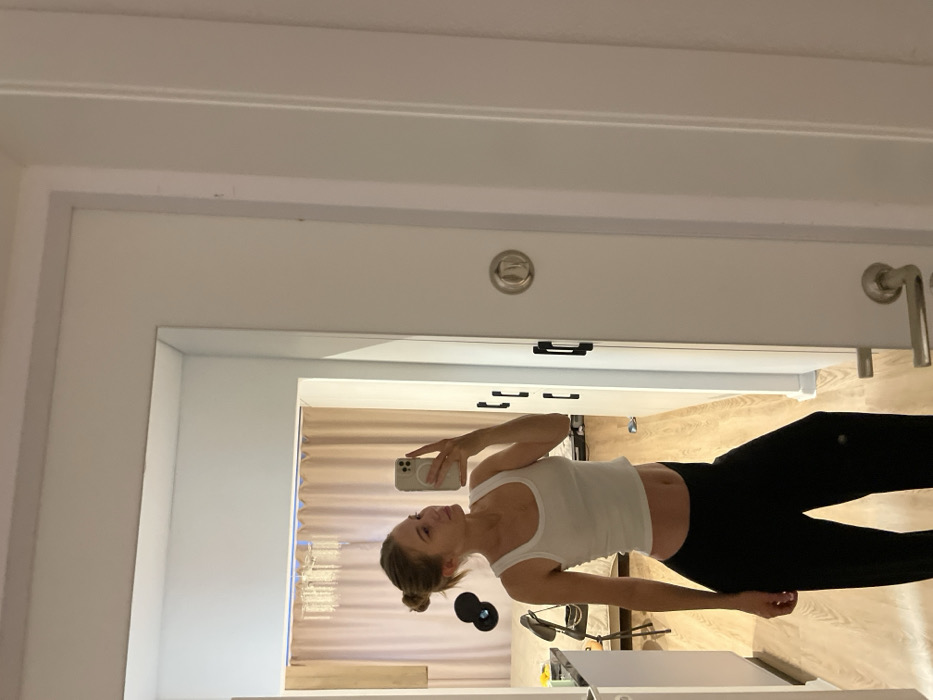 A woman is taking a mirror selfie indoors. She is wearing a white tank top and black leggings. The background shows a room with curtains and light-colored wooden flooring. A desk with a lamp and weights can also be seen. The mirror reflects a white door frame with a circular light fixture and door handle.
