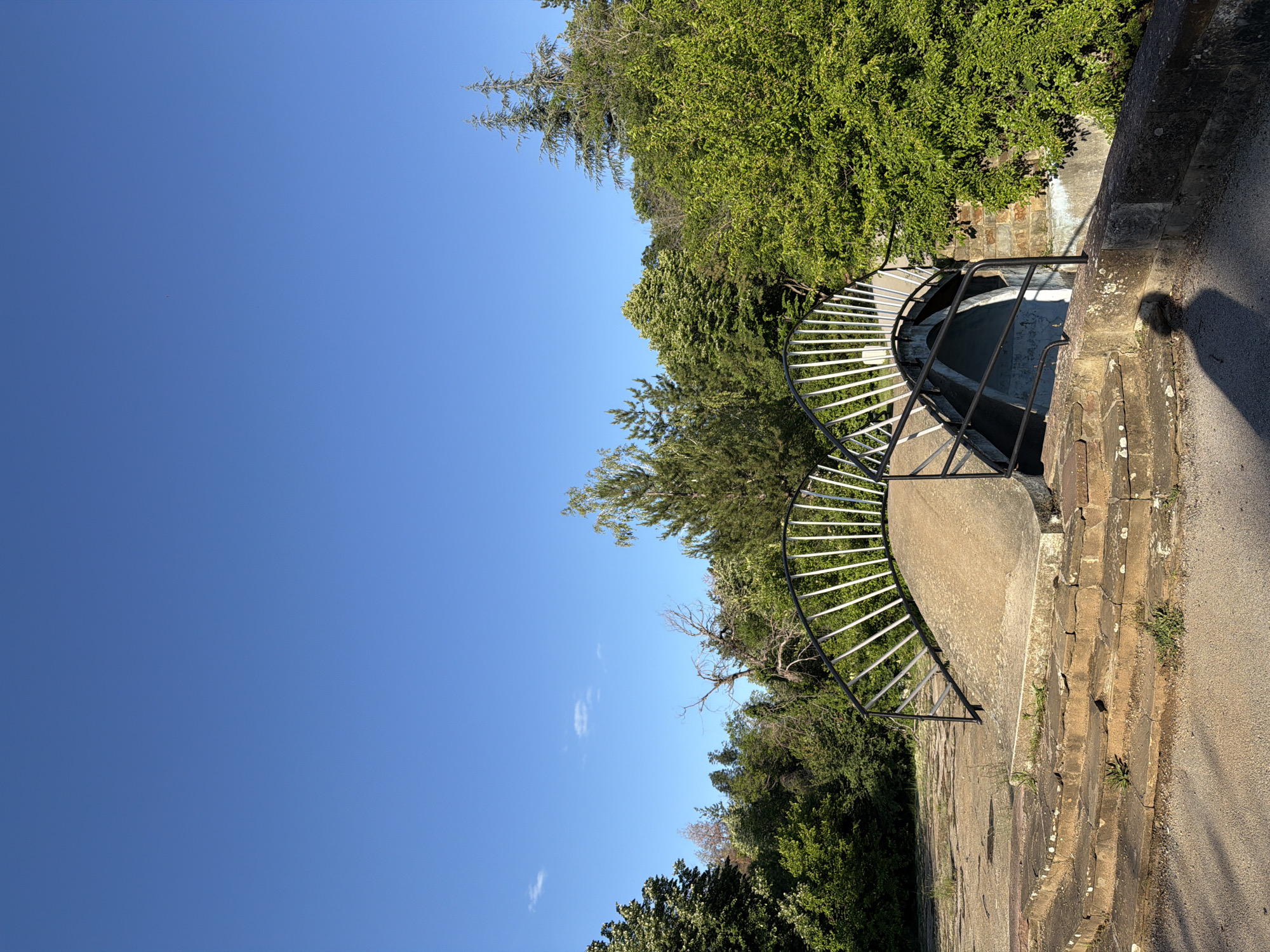 The image displays an outdoor scene under a vast, bright blue sky, occupying roughly the left half of the frame. On the right, dense green foliage and numerous trees create a vibrant backdrop, with some taller, conifer-like trees visible against the clear sky.
In the foreground and mid-ground, a distinctive circular architectural feature is prominent. It features a dark metal railing with several thin, white vertical bars, forming a semi-circle that appears to curve downwards. This structure likely covers or provides access to a deep well, cistern, or subterranean opening, as a dark, shadowed basin is visible at its base. The ground immediately surrounding this circular feature consists of weathered, light-colored stone or concrete paving, showing signs of age with irregularities and small patches of grass and weeds growing in the cracks.
To the far right, a dirt or gravel path runs along the edge of the scene, with distinct shadows cast upon it. The strong sunlight, indicated by these shadows and the clear sky, suggests it is either morning or late afternoon.
No people are present, and no specific activities are taking place, lending a peaceful and serene atmosphere to the scene. There is no visible text in the image. This tranquil setting is located in Lovech, Bulgaria, possibly within a park, garden, or historical site.