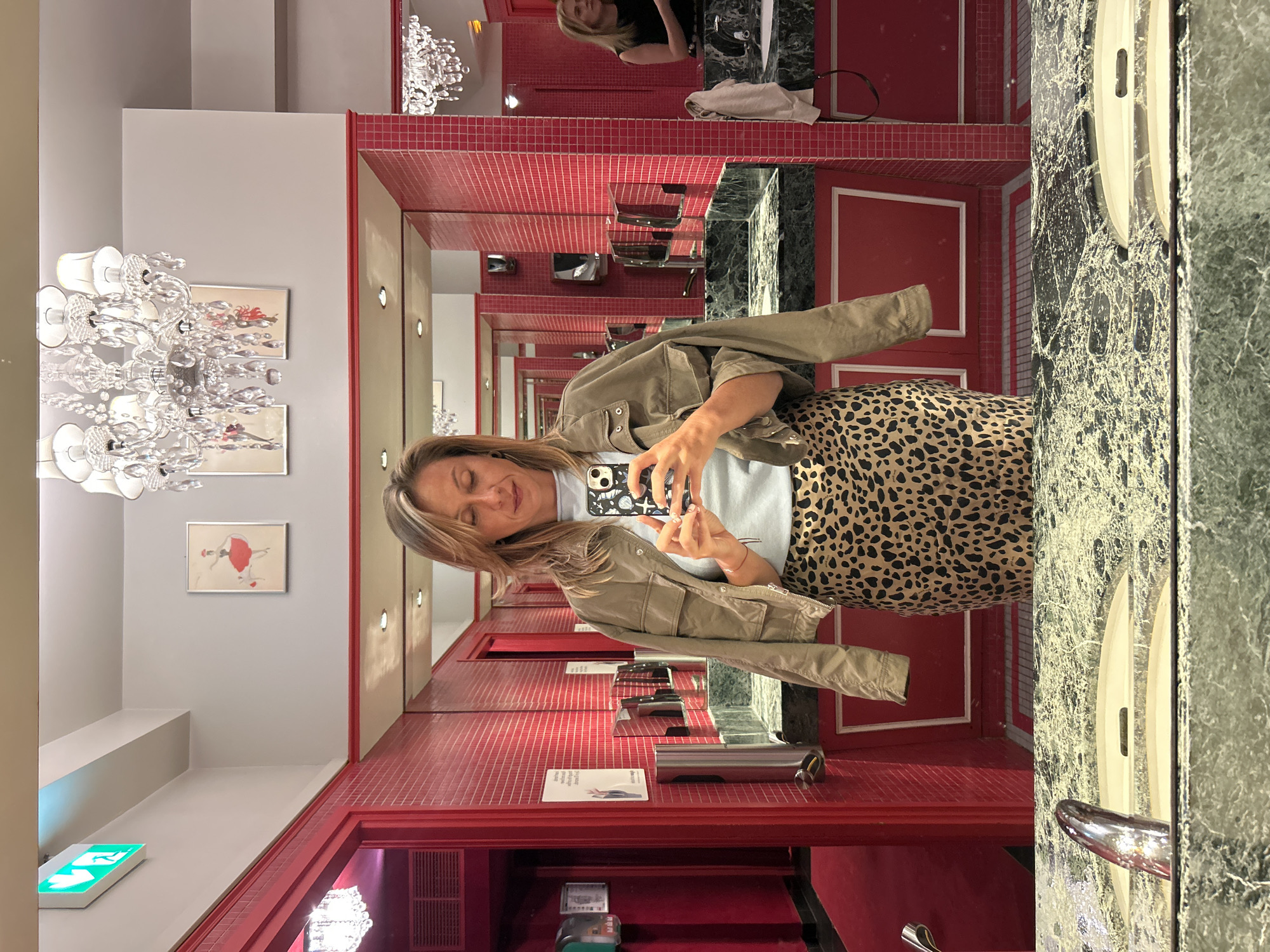 A woman stands in a well-lit, red-tiled bathroom, taking a mirror selfie. She is wearing a light blue t-shirt, a tan utility jacket, and a leopard-print skirt. She holds a smartphone in her right hand, angled to capture her reflection. The bathroom features multiple mirrors, decorative chandeliers, and framed artwork depicting ballerinas. In the background, another woman can be seen in a reflection. The overall aesthetic suggests a chic, possibly Parisian, establishment.