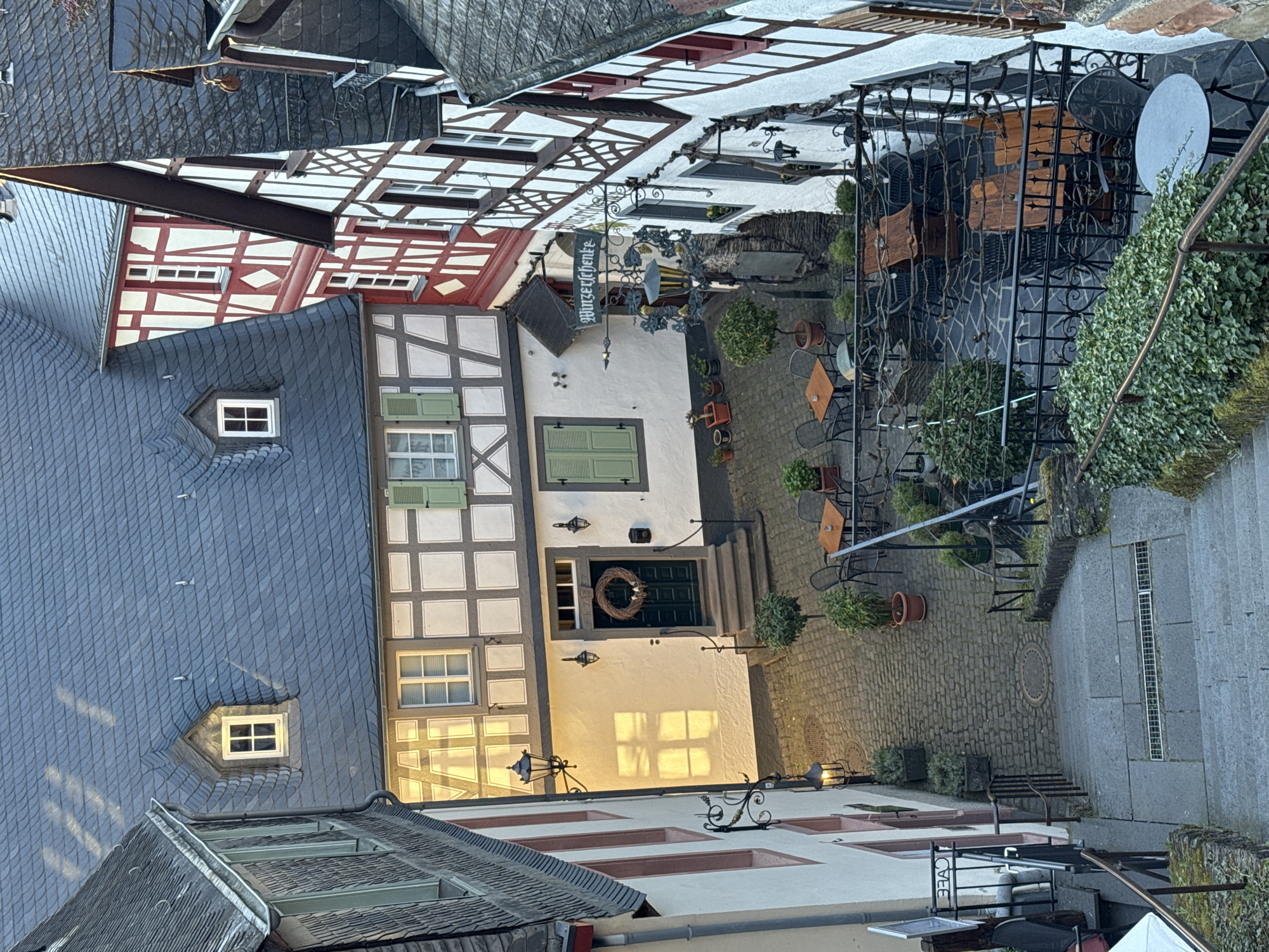 An elevated view of a street scene in Beilstein, Germany, shows multiple buildings with distinctive half-timbered facades, some adorned with red accents. A building with a dark blue slate roof is in the left foreground, punctuated by two dormer windows. Adjacent is another building with a lighter facade, featuring green shutters and a wreath on its door. A sign that reads "Winzergenke" hangs from the front of this building. Paved outdoor dining area, complete with tables, chairs, and potted plants, is in front of the building. The area is covered with decorative wrought iron fixtures. Stairs lead down to the right. The sky is not visible.