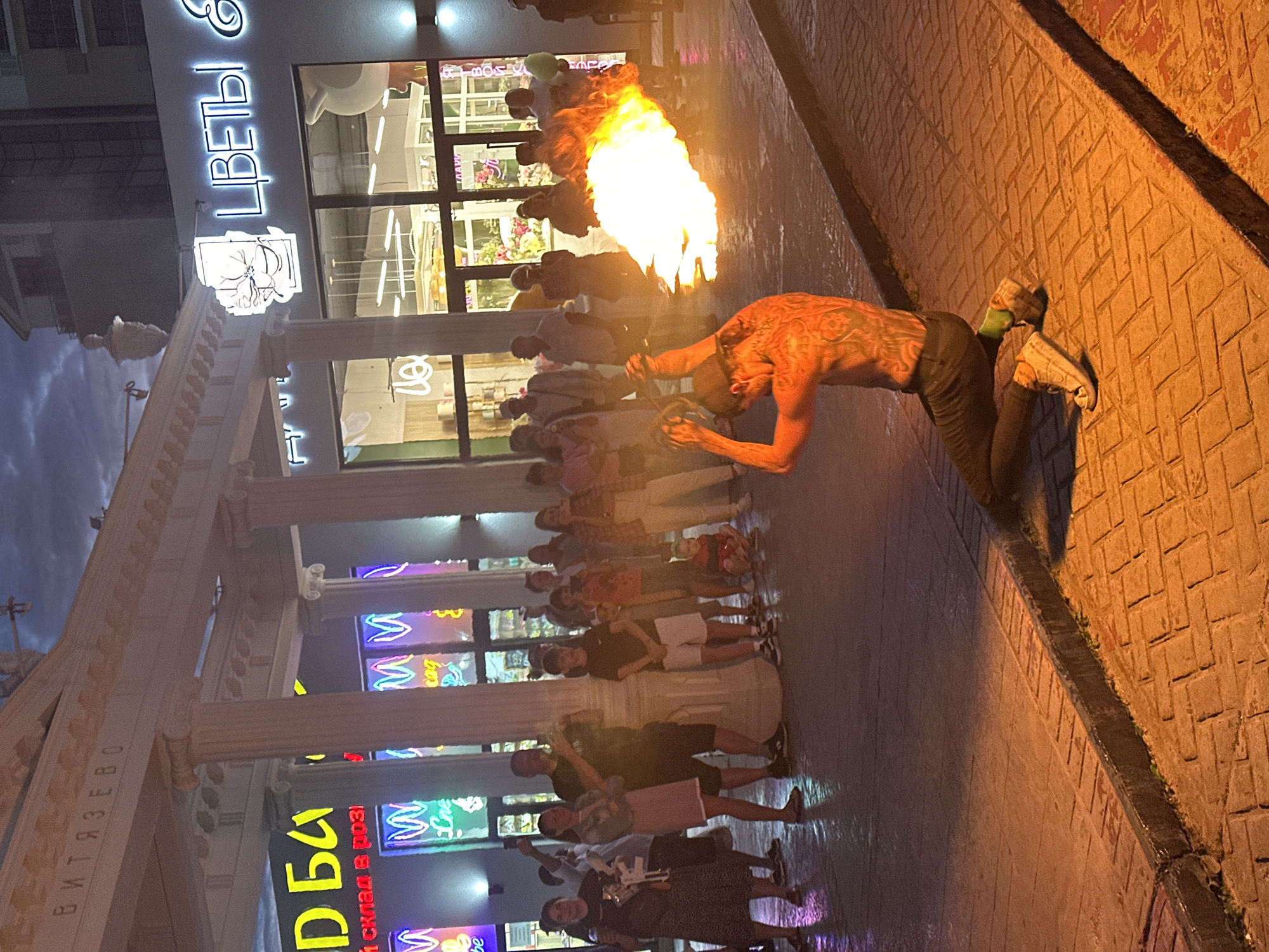 This image depicts a street scene in Vityazevo, Russia, likely during the evening, as evidenced by the twilight sky and illuminated storefronts. In the foreground, a man with extensive tattoos on his back is performing a fire-breathing act, expelling a large plume of flame from his mouth. He is kneeling on a cobblestone pavement, dressed in dark pants and sneakers. A crowd of onlookers stands behind him, observing the performance. Many individuals in the crowd are holding up their phones, presumably to record the spectacle.
The background features a row of buildings with illuminated signs. One sign, in bright neon letters, reads "ЦВЕТЫ &" (Flowers &). Another sign, partially visible, displays "Д БА" in large yellow letters, suggesting a commercial establishment. Architectural elements like white columns and a classical-style facade are also visible on the buildings. The ground is wet and reflective, indicating recent rain or a damp surface. The overall atmosphere is one of a lively public gathering, possibly a tourist area, with entertainment provided for passersby.