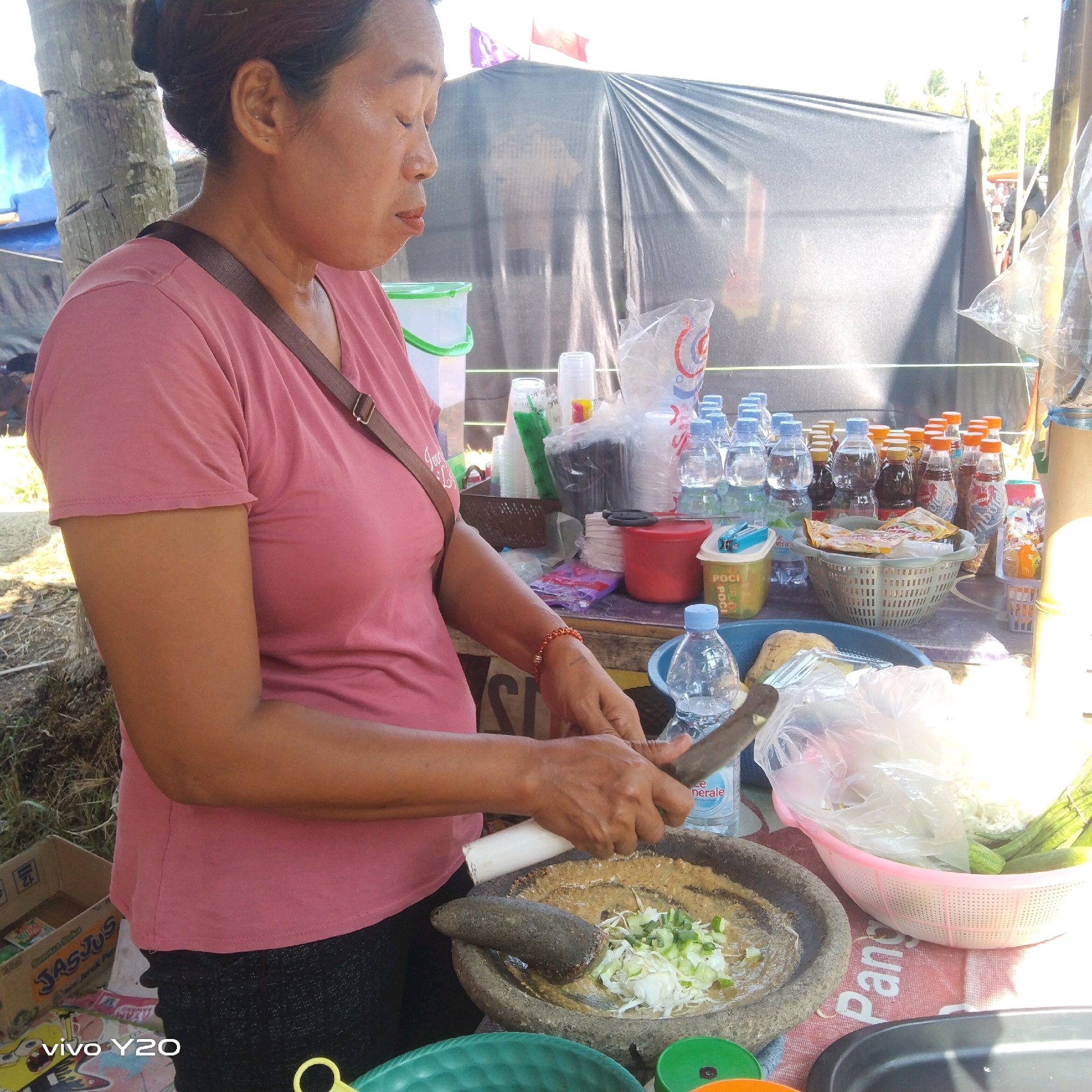 In Karawang, Indonesia, a woman is shown on a sunny day preparing food. She is standing behind a makeshift market stall, characterized by a black tarpaulin backdrop and a wooden table laden with various items. The woman, wearing a pink t-shirt and black pants, is actively engaged in grinding ingredients in a mortar and pestle. Chopped vegetables, likely for a salad or condiment, are visible within the mortar.
The table displays a collection of bottled drinks, including water and soda, as well as plastic cups and bags of snacks, suggesting she is a vendor at a local market or event. A pink basket filled with fresh vegetables, including what appear to be green beans and cucumbers, sits to the right of the mortar. The overall setting suggests an outdoor, informal commercial environment. The presence of a tree trunk and dry grass in the background hints at a rural or semi-rural location within Karawang.