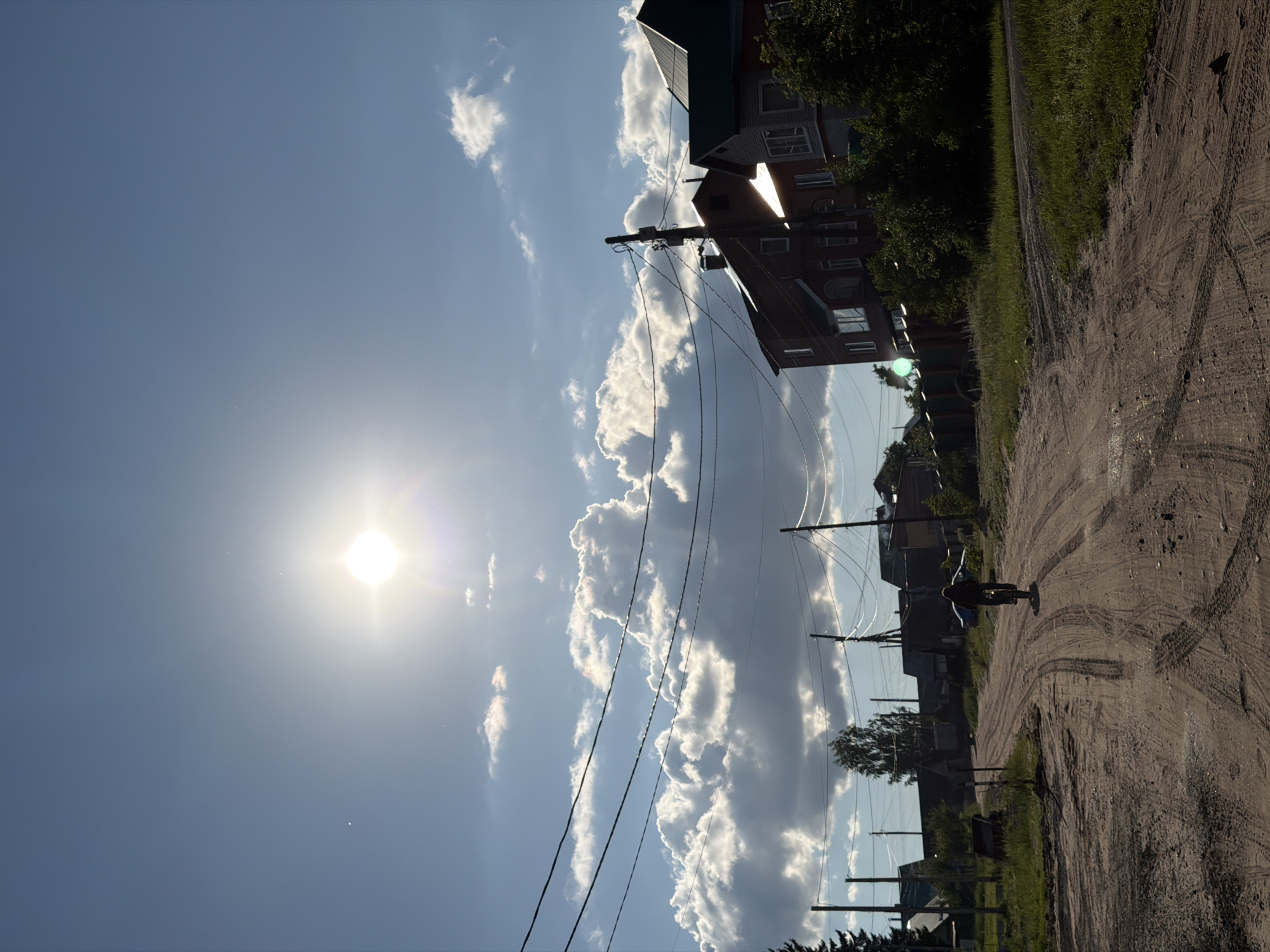 The image depicts a sun-drenched, rural street scene. In the foreground, a person is riding a bicycle on a dirt road, which is marked with tire tracks. The road is lined with grass and some bushes on the right side. In the background, there are several residential buildings, some appearing to be two-story homes with dark roofs. Power lines are strung across the sky, supported by utility poles. The sky is bright blue with large, fluffy white clouds and the sun is prominently visible, casting a bright glare. The overall impression is that of a quiet, sunny day in a village or town.