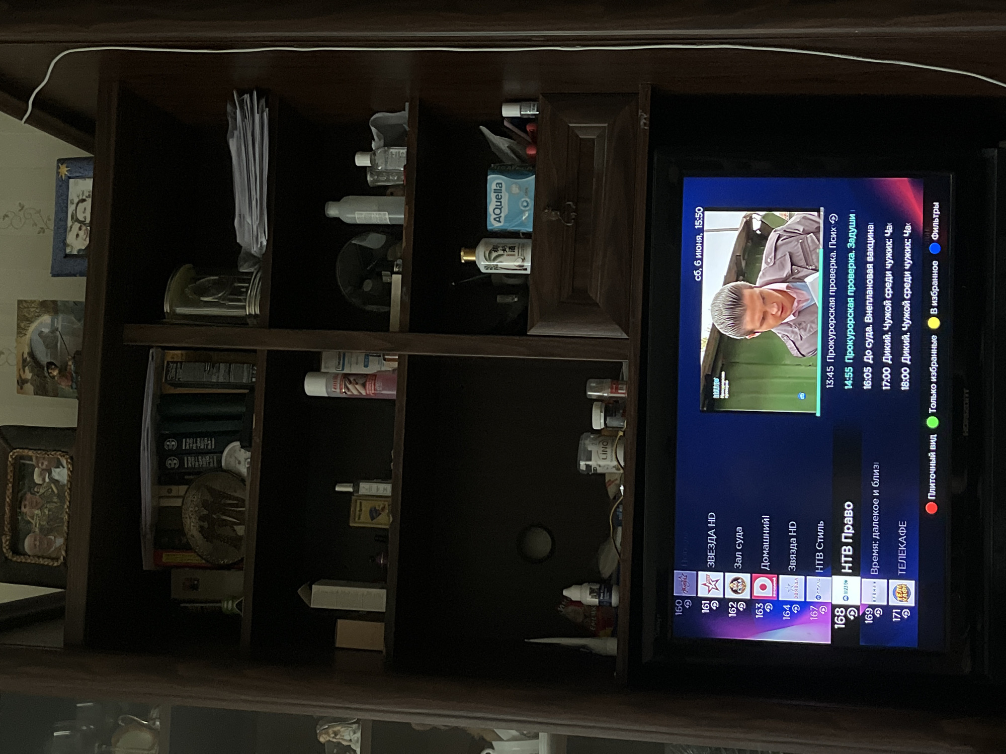The image depicts an interior scene featuring a dark wooden shelving unit or cabinet with a television positioned centrally at its base.
The uppermost shelf displays three framed photographs. The largest, on the left, shows three individuals: an older man, a man in military fatigues, and a woman. To its right, a smaller blue framed photograph depicts two individuals, likely a couple. A third, indistinct framed image is further to the right.
The second shelf from the top holds a silver miniature Eiffel Tower, a dark circular decorative plate or medallion with etched figures, and several books. To the right, a stack of white papers rests beside a clear glass object, with more books visible behind them.
The third shelf contains two tall, light-colored rectangular boxes, a small golden-colored bottle or container, and other miscellaneous bottles and personal care items.
The shelf directly above a central wooden drawer features a white squeeze tube, a partially visible snack bag, a small white bottle labeled "Pet Care" (partially obscured), a pack of "AQuella" wet wipes, a white bottle with a gold cap, and red earbuds. A small white rollerball bottle is also present on the far right.
The television, a "HORIZONT" brand model, displays an electronic program guide (EPG) in Russian. Channel 168, "НТВ Право" (NTV Law), is highlighted. The display shows "сб, 6 июня, 15:50" (Saturday, June 6, 15:50). A live video feed on the right side of the screen depicts a male individual with slicked-back hair, wearing a light-colored jacket, looking downwards or to the side against a green, possibly outdoor or industrial background. Program listings include "Прокурорская проверка. Псих" (Prosecutor's Check. Psycho), "До суда. Внеплановая вакцина" (Before the Court. Unscheduled Vaccine), and "Дикий. Чужой среди чужих" (Wild. Stranger Among Strangers). Interaction options like "Плиточный вид" (Tiled View) and "В избранное" (Add to Favorites) are visible at the bottom of the screen.
A white cable runs vertically along the right side of the cabinet. The room appears dimly lit.
Given the prominence of a "HORIZONT" brand television, a Belarusian electronics manufacturer headquartered in Minsk, and the Russian language interface, the scene is located in Minsk, Belarus.