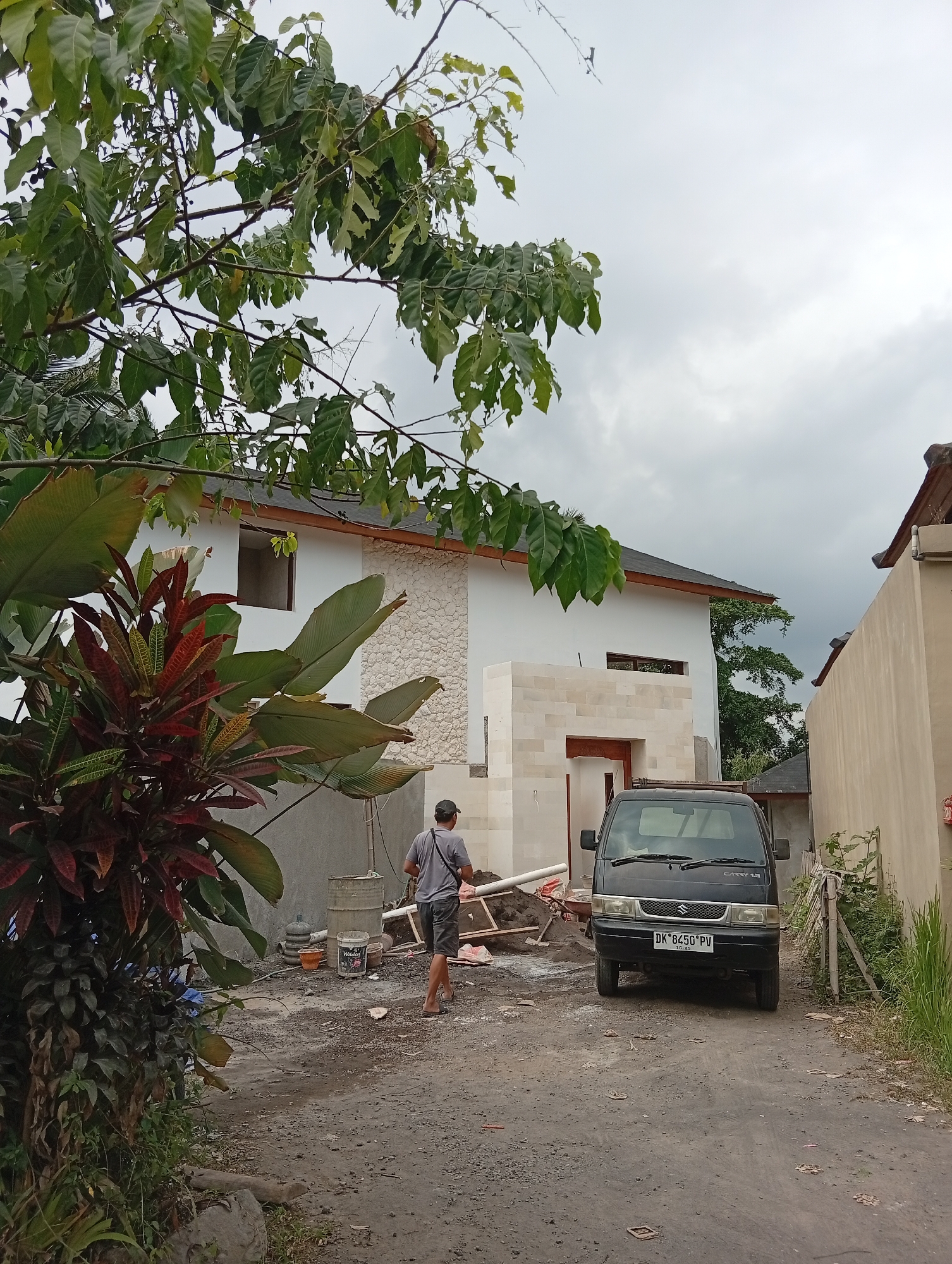 The image captures a construction site in Ubud District, Indonesia, on a cloudy day. A black Suzuki Carry van, with the license plate DK 8450 PV, is parked on a dirt road. To the left of the van, a partially constructed white building with stone accents is visible. Several construction materials, including buckets, piping, and piles of dirt, are scattered around the site. A man wearing a grey t-shirt and shorts walks away from the camera, heading towards the building. Lush green foliage, including large-leafed tropical plants and a tree with broad leaves, frames the scene, with some branches extending into the upper part of the image. The overall impression is one of ongoing construction in a tropical environment.