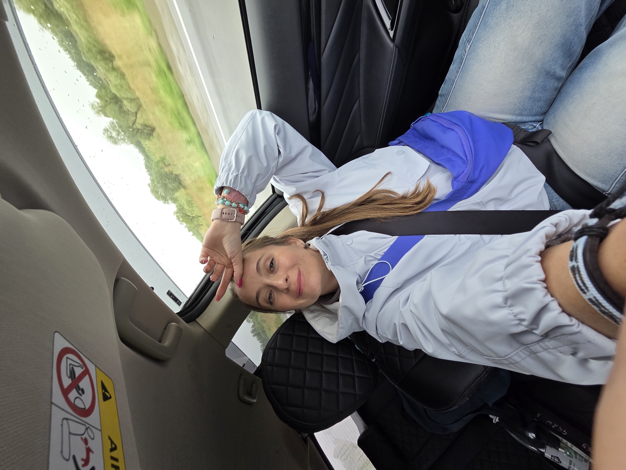 A young woman is captured lying on her back across the rear seats of a car, looking directly at the camera with a gentle smile. She has light brown hair and is wearing a light grey or light blue hooded jacket, blue jeans, and a bright blue fanny pack/sling bag strapped across her. Her left hand is raised to her forehead, revealing pink nail polish, a light pink smartwatch, and several bracelets on her wrist (some beaded, some with blue accents).
The car's interior features dark, possibly quilted, upholstery on the seats and headrest, contrasting with the lighter beige or grey color of the interior ceiling and door panel. A black seatbelt is visible across her torso.
Through the car window, a blurred green landscape of trees and what appears to be a road or field can be seen, indicating the vehicle is in motion. Numerous raindrops are visible on the window, suggesting rainy weather or recent rainfall. The scene is brightly lit, consistent with daytime.
Notable details within the car include a beige grab handle above the window and a partially visible warning label on the car's interior pillar. The label features a red circle with a diagonal line over an icon depicting a child in a rear-facing car seat, and a yellow triangle with an exclamation mark alongside the word "AIRBAG."
The overall impression is a relaxed moment during a car journey in Бабстово, Russia.