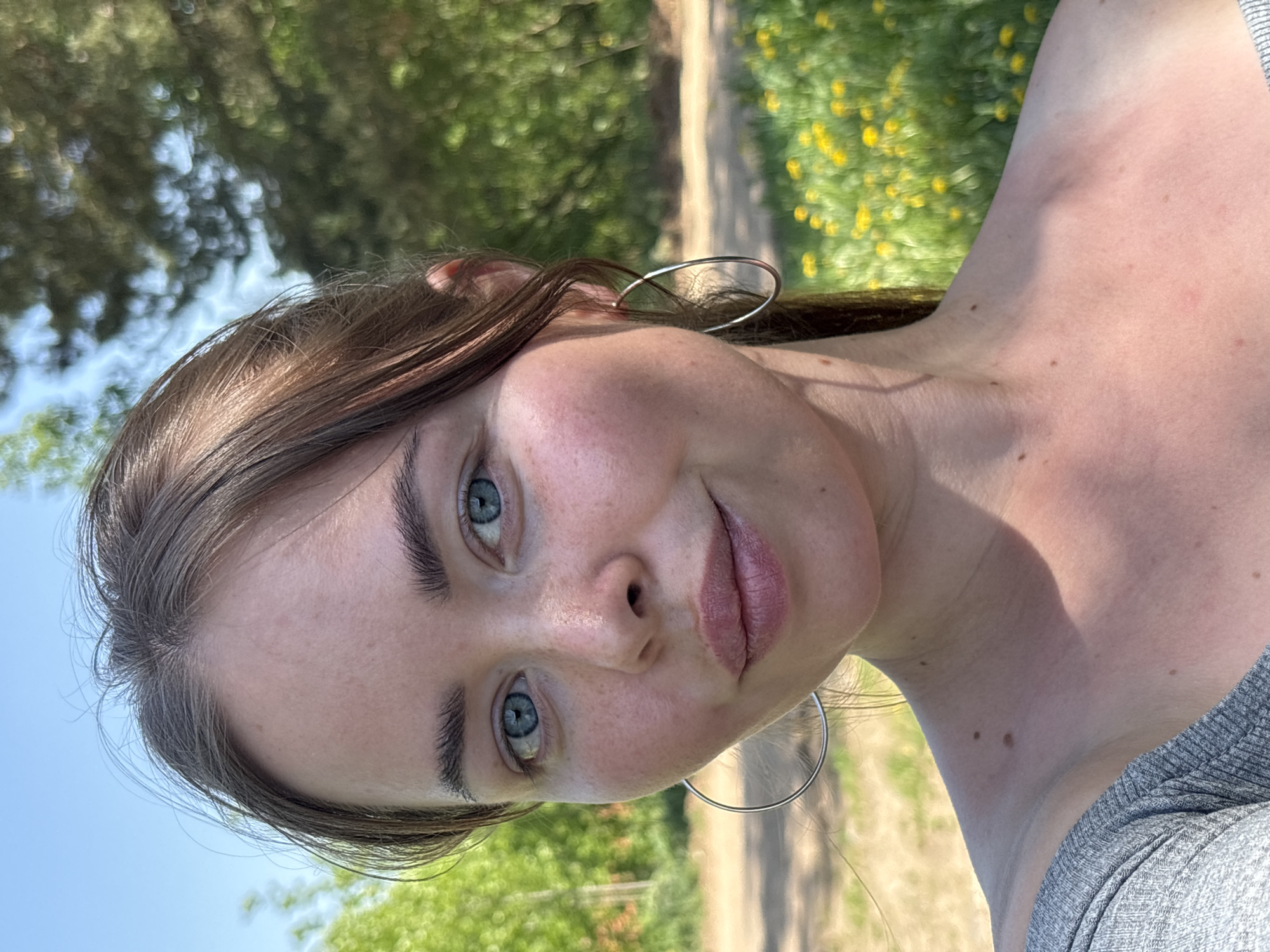 A selfie of a woman with light brown hair and blue eyes, wearing silver hoop earrings. The background appears to be outdoors, with trees, flowers, and a dirt path visible. Location: Unknown, not verifiable as Русский Вожой, Russia.