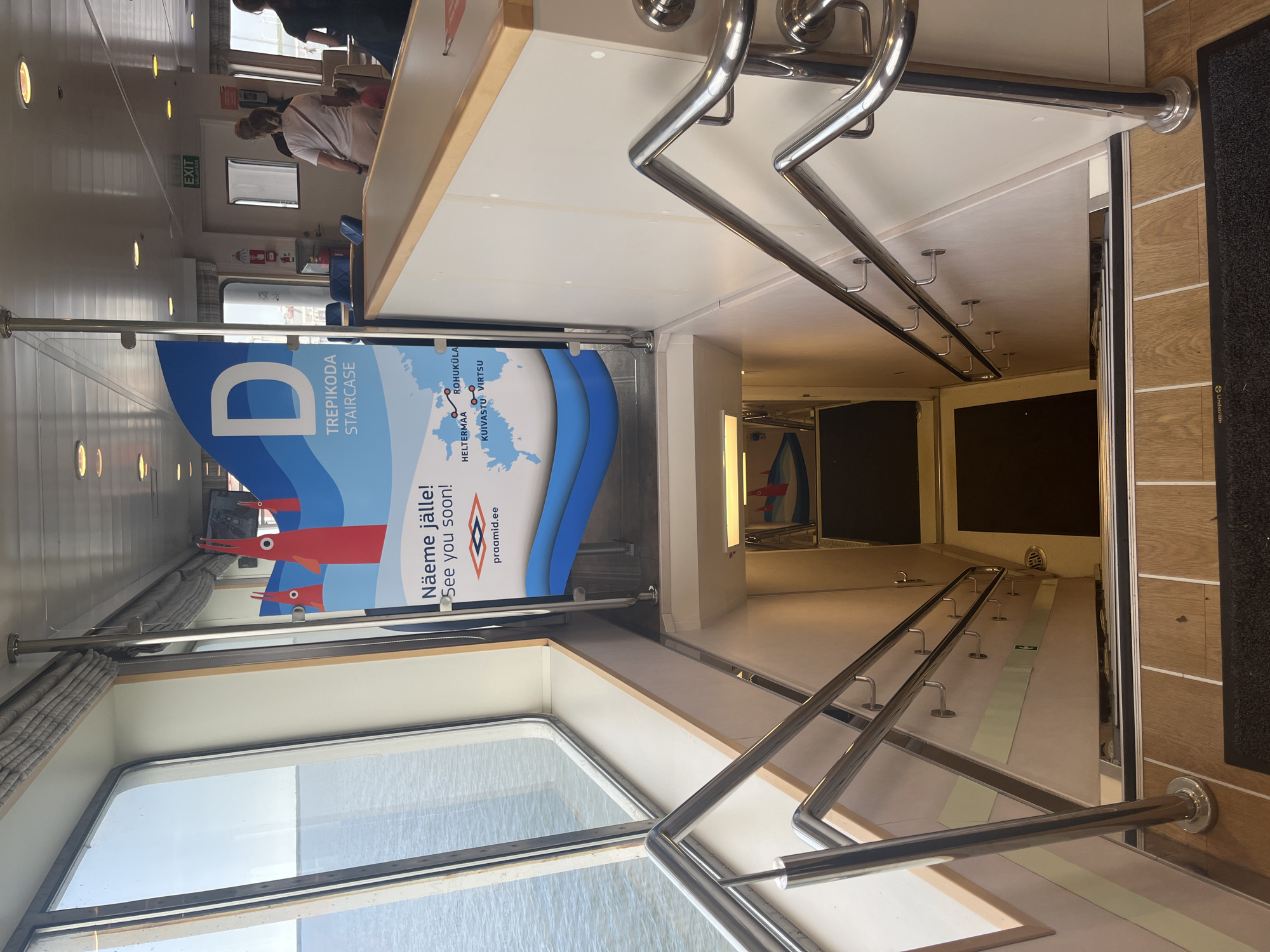 The image shows a downward view of a staircase on a ferry, likely docked at Kuivastu, Estonia, based on the map shown on the adjacent sign. The staircase has polished metal railings on both sides and wooden steps with black non-slip treads. The walls of the stairwell are beige, with decorative metallic accents and visible hooks. At the bottom of the visible stairs, a doorway is visible, with a framed picture of red cartoon-like figures on the wall to the left.
The sign next to the staircase features a large blue "D" and the text "TREPIKODA STAIRCASE" in English and potentially Estonian, along with a map of Estonia showing ferry routes, including Kuivastu and Virtsu. Below this, the text "Näeme jälle! See you soon!" and the website "praamid.ee" are displayed.
In the background, on the upper deck, there are people dressed casually. An "EXIT" sign is visible above a doorway. The environment suggests a public transport vessel, with the sea visible through large windows on the left side of the image.