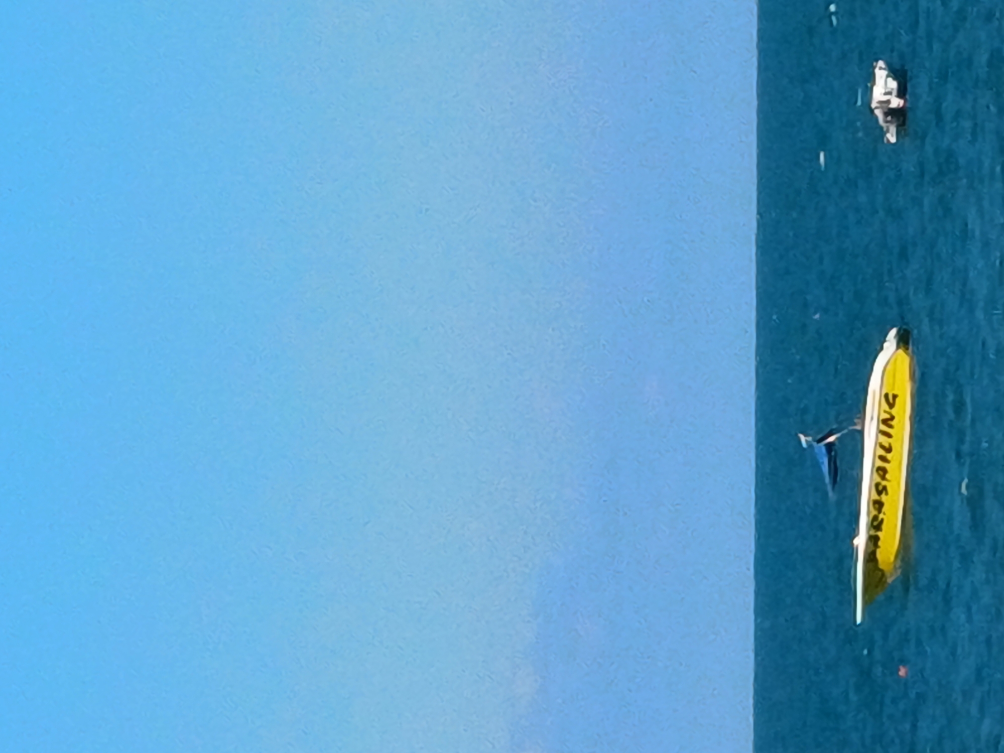 The image shows a bright blue sky and a calm, deep blue sea. Two boats are visible on the water. The boat on the left is yellow with "PARASAILING" written in black letters on its side. It has a blue flag with black and white stripes at its stern. There is also a small orange buoy in the water near this boat. The boat on the right is smaller and white, appearing to be a motorboat. The horizon line is visible where the sky meets the sea. The scene suggests a clear day at a coastal location, possibly a resort area given the presence of a parasailing boat. It is not possible to determine the specific location within Sochi, Russia, from the provided image.