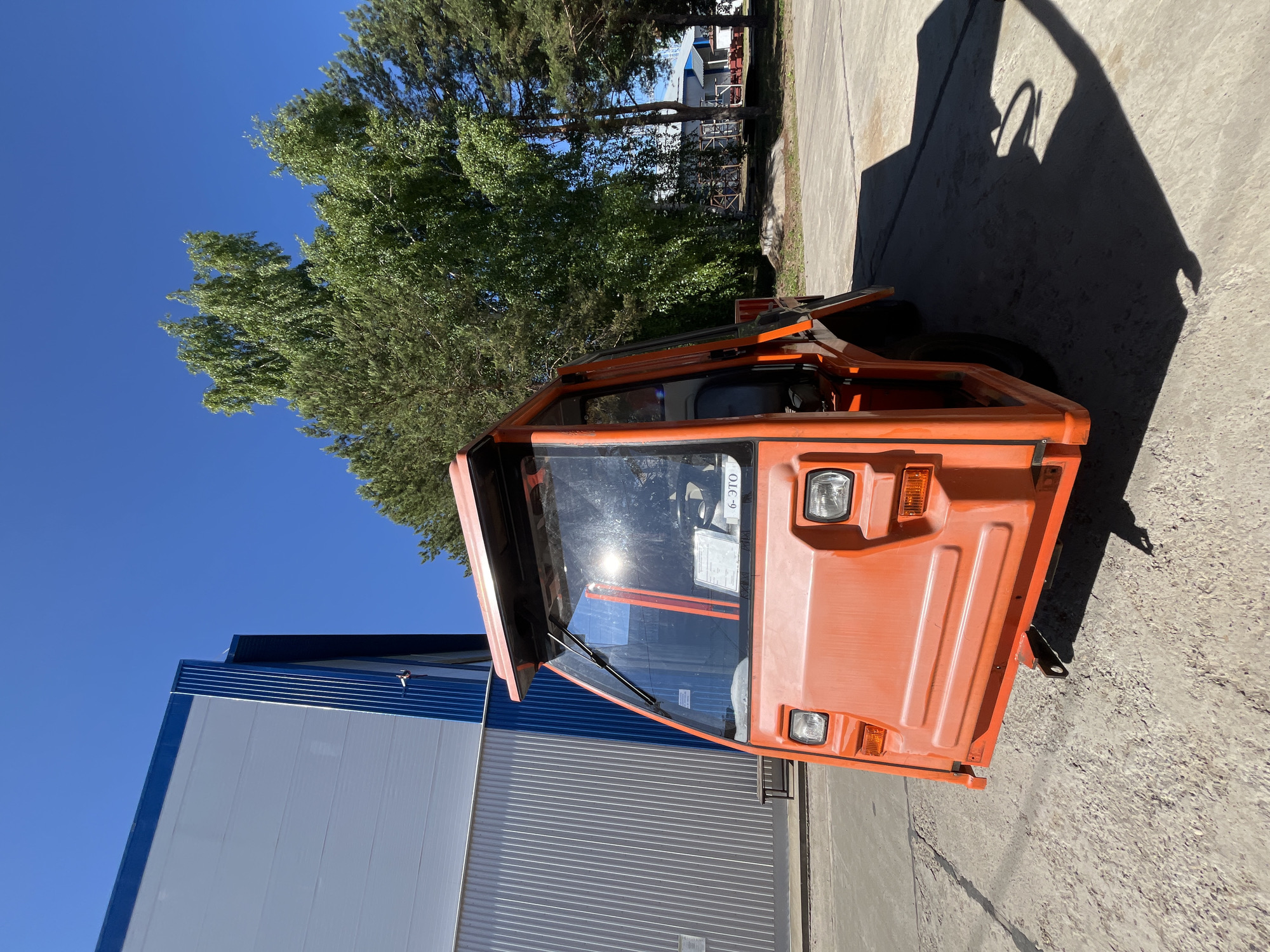 An orange industrial utility vehicle is parked on a concrete surface in front of a white and blue building under a clear blue sky. The vehicle has a prominent windshield with a wiper, two headlights, and orange side marker lights. There is a visible shadow cast by the vehicle on the ground. To the right of the vehicle, several green trees are visible. The building has vertical silver ribbed paneling on its lower section and white paneling on its upper section, bordered by a blue trim. The scene appears to be outdoors in a daylight setting.