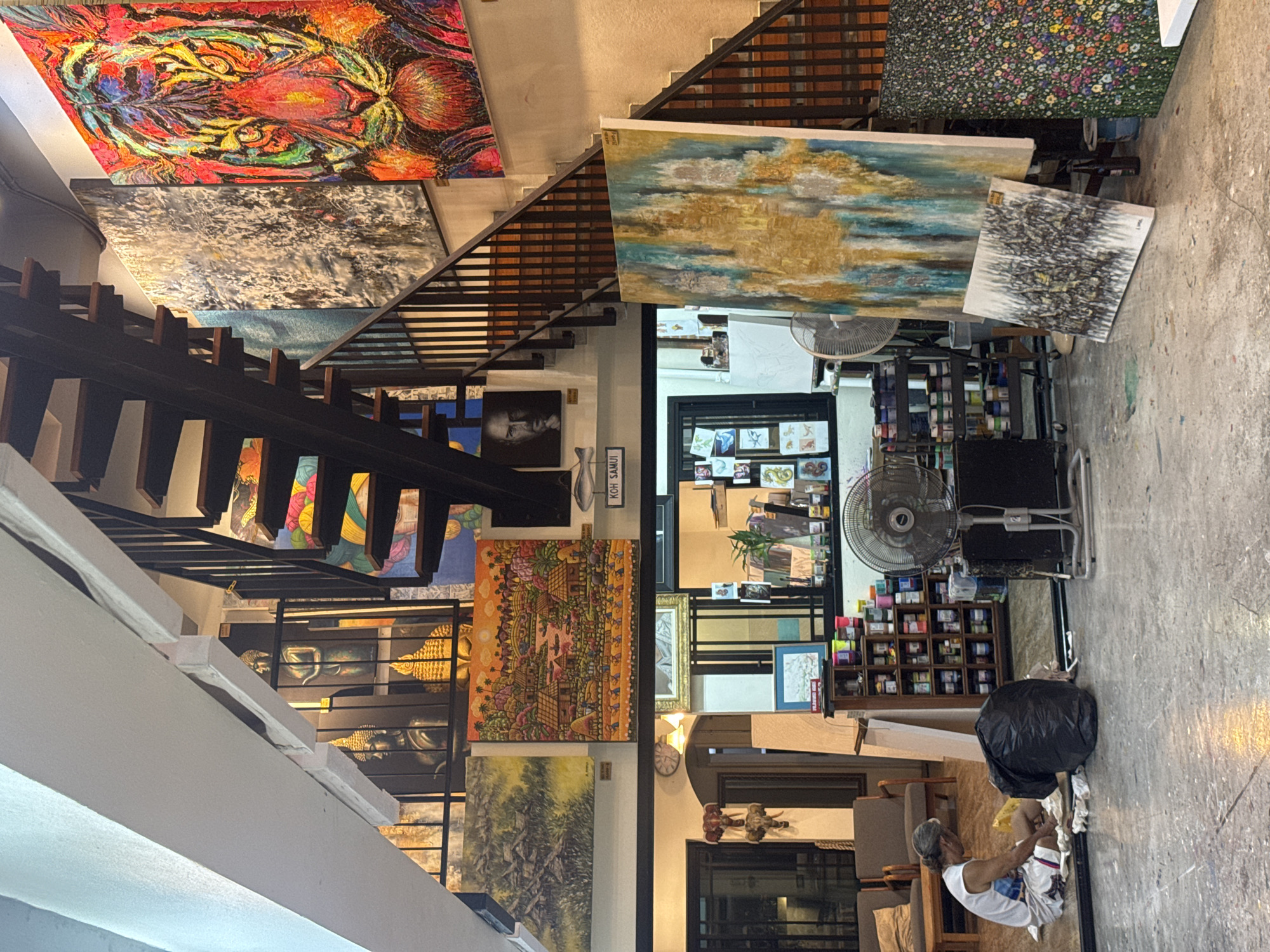 The scene depicts the interior of an art studio or gallery in Baan Bo Phut, Thailand. The space is characterized by a staircase with a dark railing dividing the room, multiple paintings of various sizes and styles displayed on the walls, shelves filled with art supplies, and two electric fans. A man is sitting on the floor near a garbage bag. The overall environment suggests a workspace dedicated to artistic creation and display.