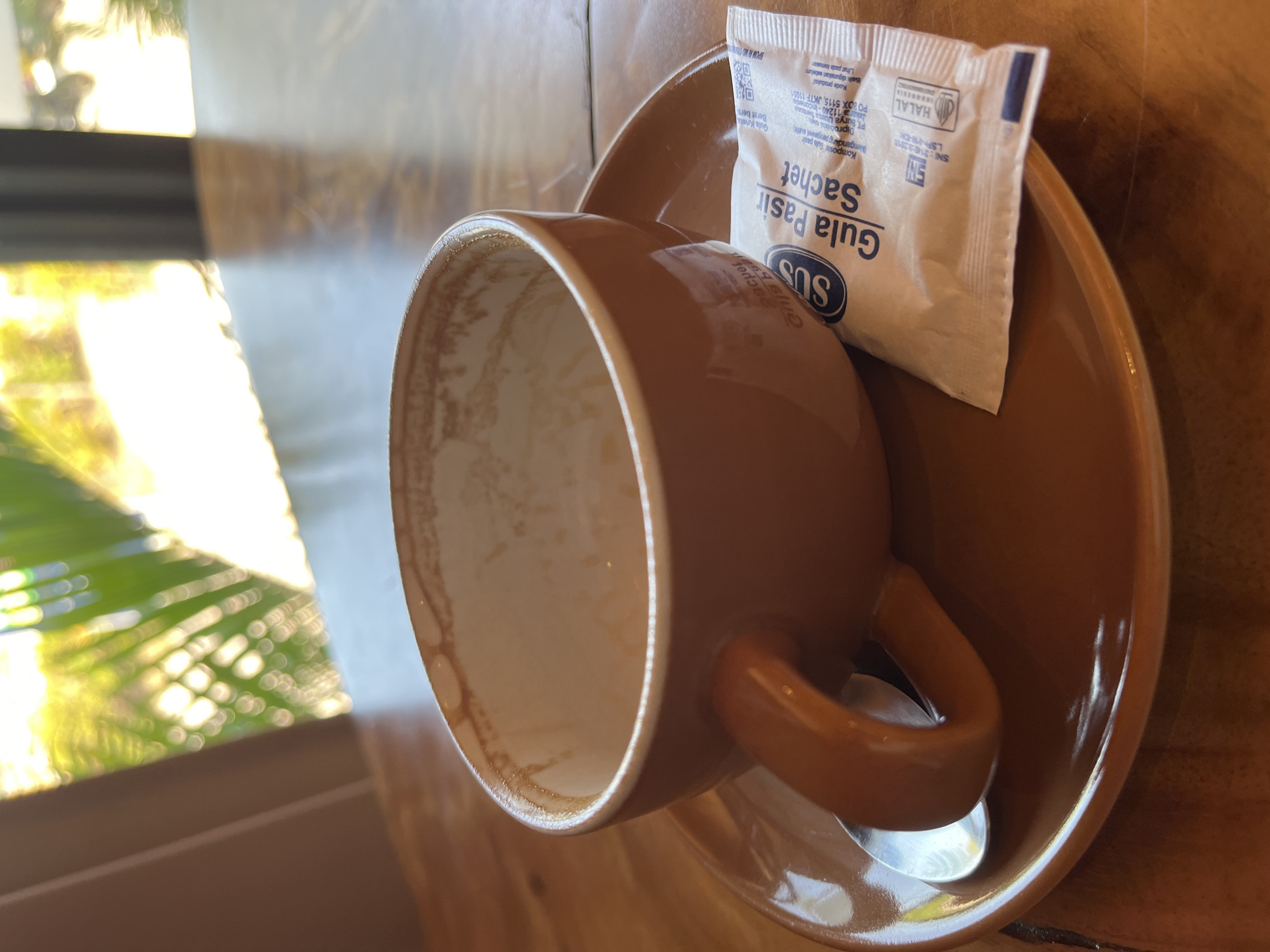 An overhead, close-up shot shows an empty, brown coffee cup and saucer on a wooden table. A silver spoon rests on the saucer to the left of the cup, and a small, white packet of sugar with blue text sits to the right of the cup. The sugar packet has the words "Gula Pasir Sachet" printed on it. In the background, out of focus, sunlight streams through foliage, suggesting an outdoor or well-lit indoor setting. The scene is likely in Kemloko, Indonesia, given the "Gula Pasir Sachet" which is Indonesian for "sugar sachet".