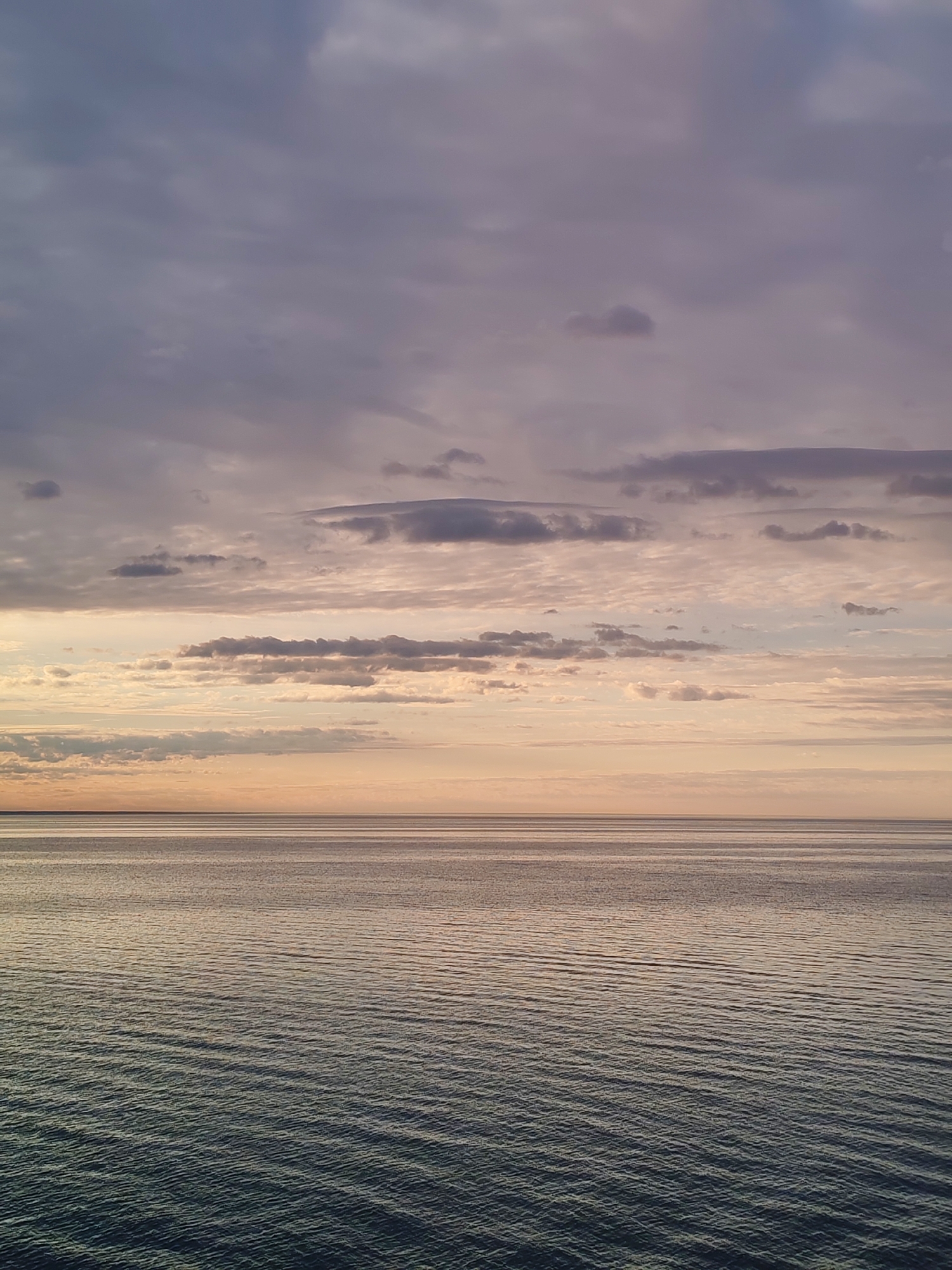 The image depicts a calm body of water under a sky filled with various cloud formations. The sky transitions from a light peach color near the horizon to darker shades of gray and purple higher up. The water is textured with subtle waves, reflecting the light and color from the sky. No people or specific objects are visible, only the natural elements of water and sky. The image is a serene landscape scene. The location is unknown and not evident from the image. Therefore, the location cannot be determined.