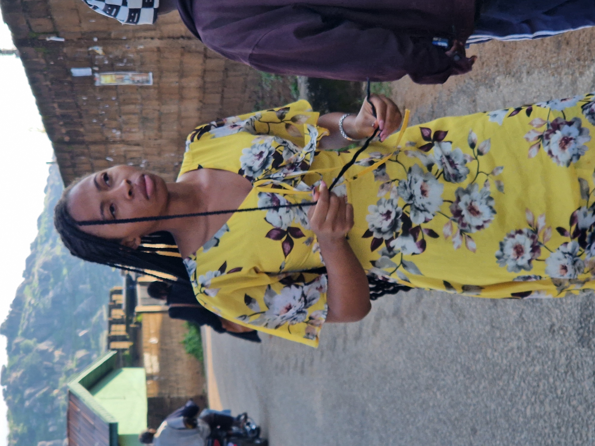 A young woman with long, dark braided hair stands on an asphalt road in Kabong, Nigeria, wearing a yellow floral dress. She is looking to her left, her expression neutral. Her right hand holds a dark string or thread, while her left hand is clasped around her right wrist, where a silver bracelet is visible. In the background, a man in a purple jacket and a checkered hat stands to the right of the frame, partially obscuring the woman. A motorcycle with a rider is visible in the distance on the left side of the road. Behind the woman, a brick wall lines the street, and beyond that, a green building with a corrugated roof is visible. Rocky hills rise in the far background. The lighting suggests it is daytime, possibly late afternoon.