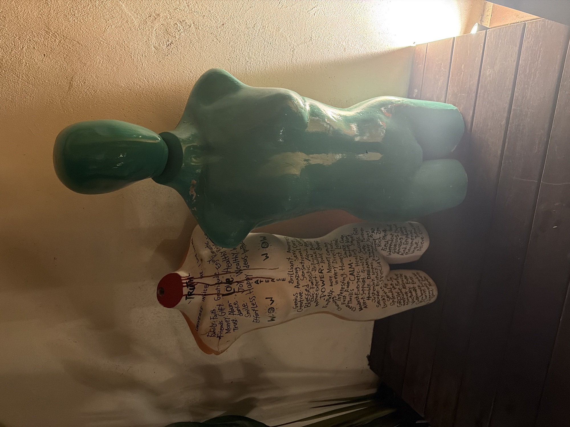 A photograph shows two mannequin torsos, one green on top of a white one with words written all over it, next to a brown wooden wall.