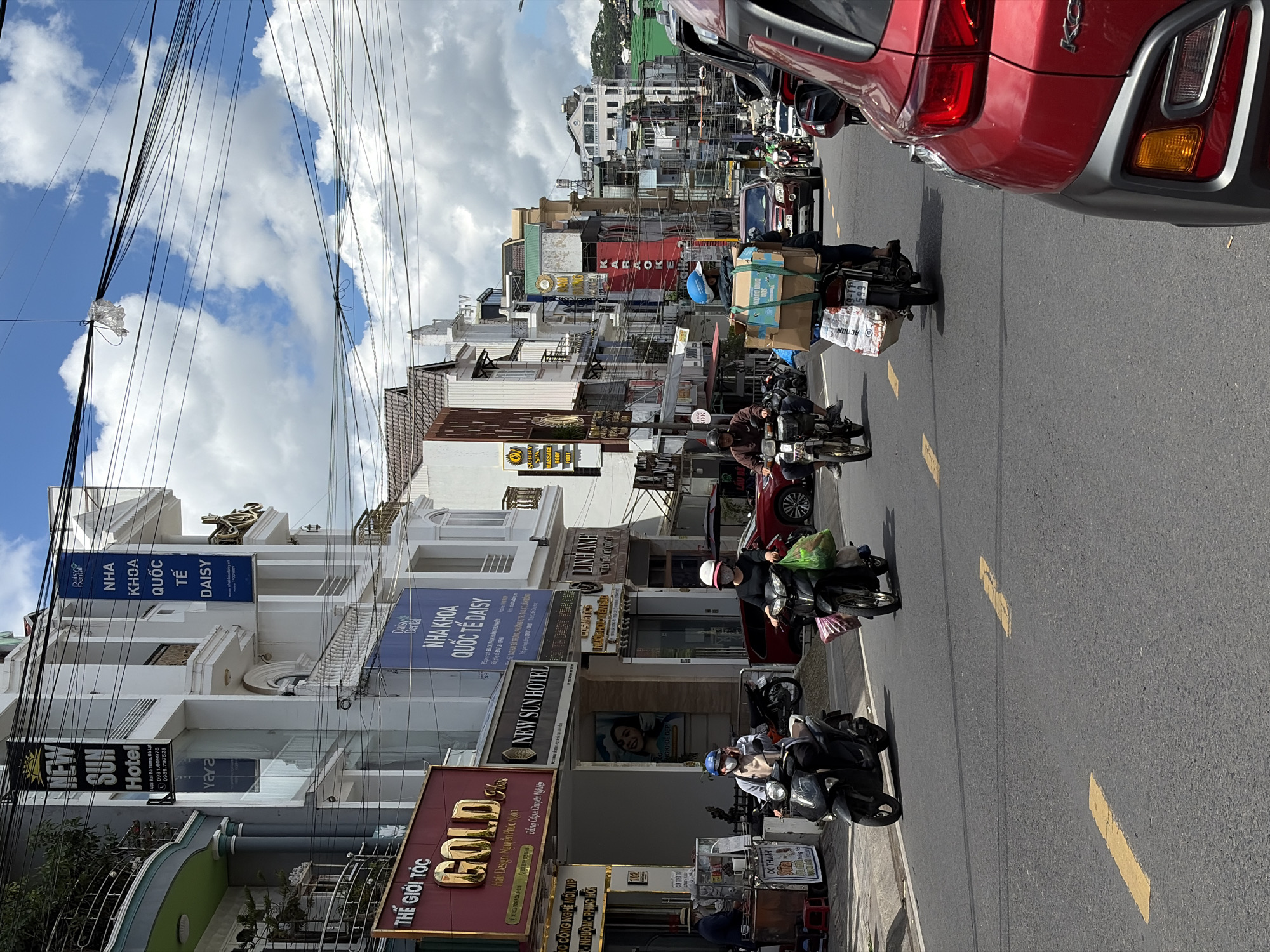 This image depicts a street scene in Da Lat, Vietnam, with a focus on the architecture and street life. The buildings lining the street are a mix of modern and slightly older structures, featuring balconies, large windows, and prominent signage. One building displays a banner for "NEW SUN HOTEL" with contact information. Another building has a large, red sign with gold lettering that reads "GOLD" and below it, "THE GIOI TOC" and "Hair Design Nguyen Phuc Ngan". Further down the street, there are signs for "NHA KHOA QUỐC TẾ DAISY".
The street itself is paved and marked with yellow lines. The sky is a mixture of blue with fluffy white clouds. Several motorbikes are present, with some individuals riding them and others parked. One motorbike in the foreground is carrying a large cardboard box and some packaged items strapped to the back. Another motorbike further back is carrying shopping bags. A red car is visible in the foreground on the right side of the image, partially obscuring the view of the street. The scene appears to be during the daytime, under bright sunlight. The overall impression is of a busy urban street in a Vietnamese city.