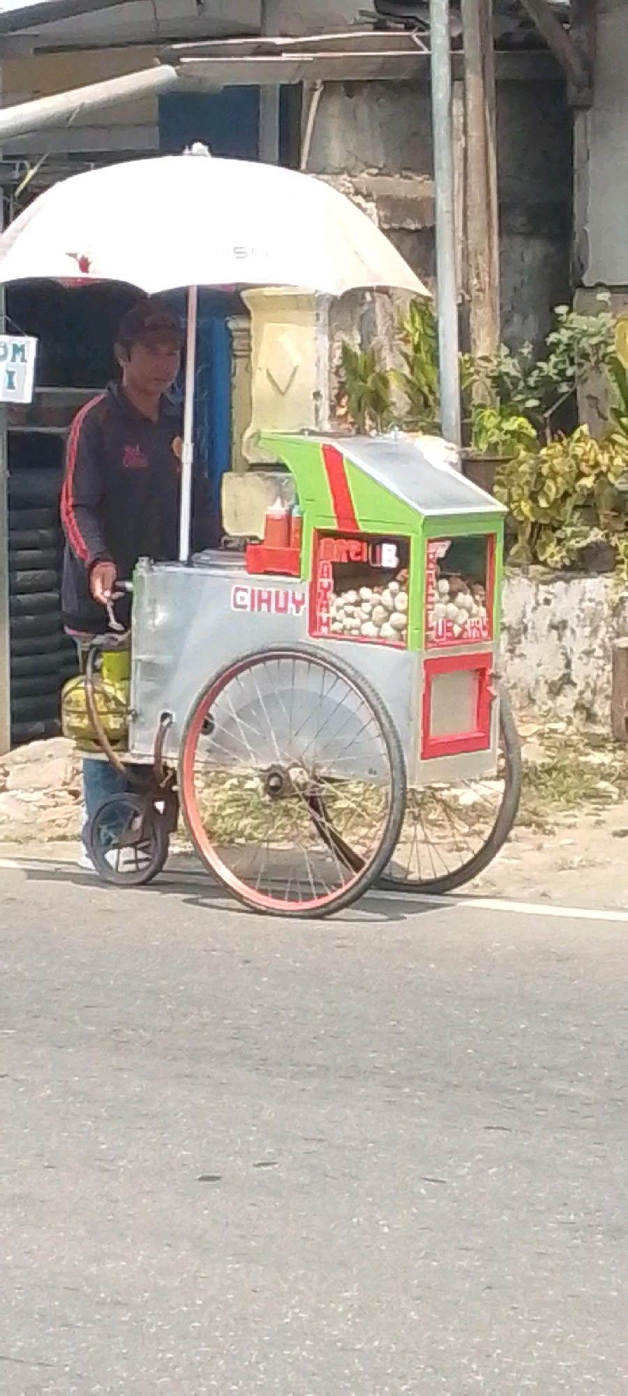 A street vendor is standing behind a mobile food cart on a paved road. The vendor, a man wearing a dark jacket with red stripes, is holding a spoon. The food cart is silver with a green and red top, and it displays the word "CIHUY" in red lettering on its side. Inside the cart, there are many round, light-colored food items, possibly meatballs or eggs, arranged in transparent compartments. A red ketchup dispenser is visible on the counter. The cart has large spoked wheels, one of which is red. A white umbrella is positioned over the cart. In the background, there is a building with a rough, textured wall and some greenery. The location is identified as Sukasari, Indonesia.