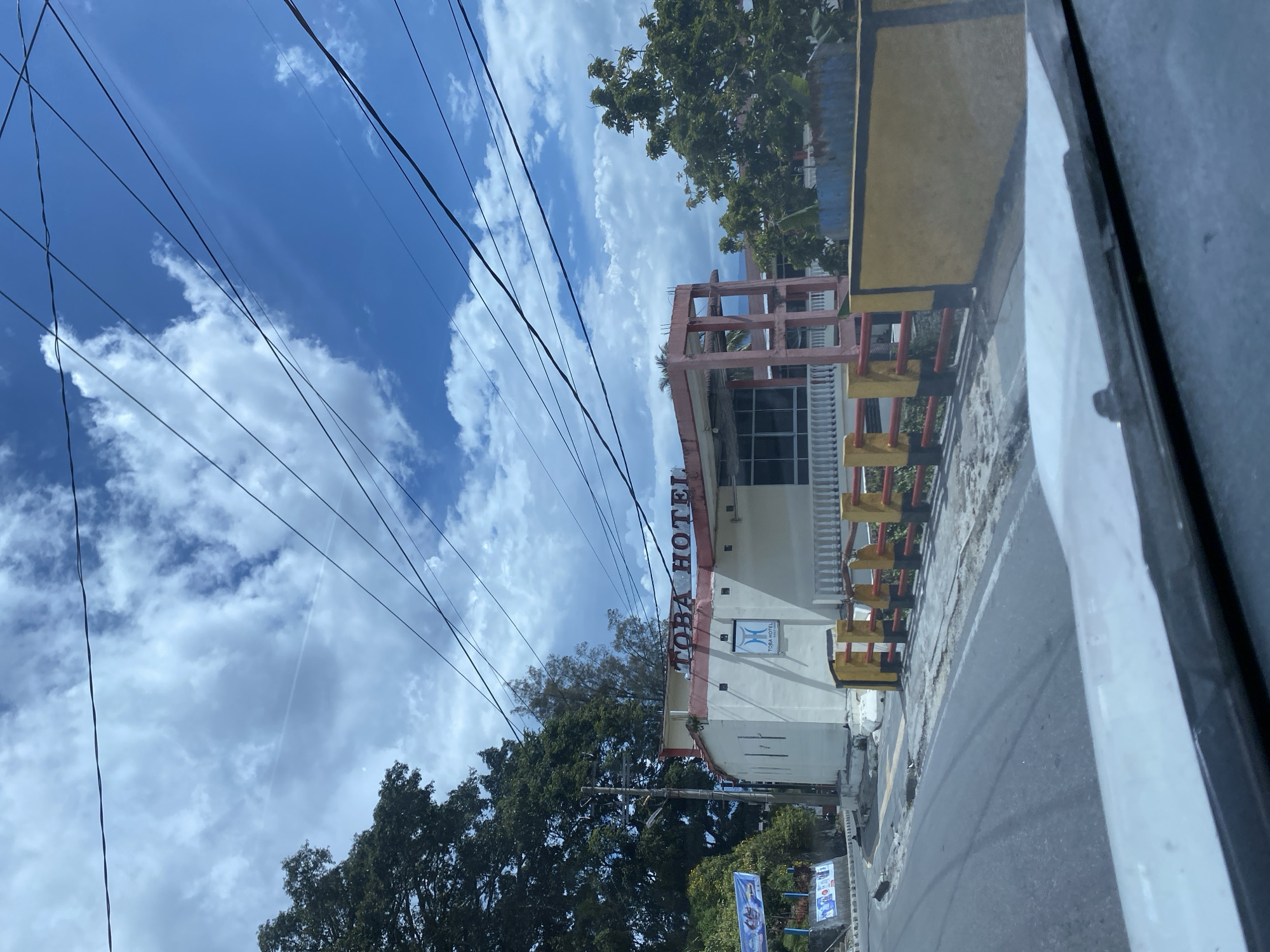 The image shows a street view in Parapat, Indonesia, featuring the "TOBA HOTEL." A white building with red accents is visible, displaying the hotel's name in red lettering on the facade. A smaller logo for Toba Hotel is also present on the building. A white railing with red and yellow supports lines the walkway in front of the hotel. Trees and a blue sky with white clouds are in the background, along with overhead power lines. The street in the foreground appears to be asphalt and shows some signs of wear.