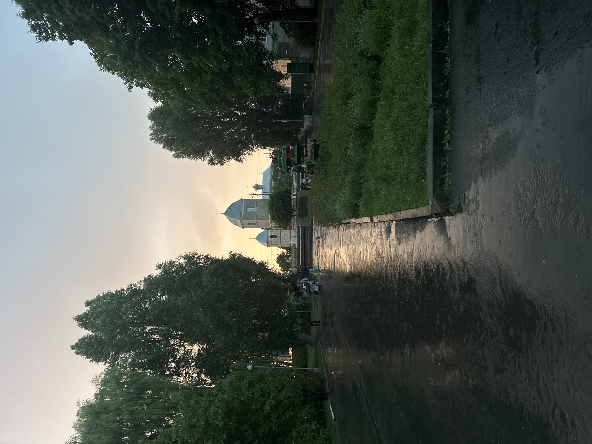 The image depicts a view from a wet, paved walkway in Ternopil, Ukraine, likely at dusk. The sky is a soft gradient of pale orange and light blue, suggesting either sunrise or sunset. Several mature trees with dense green foliage line the left and right sides of the walkway, framing a central view. In the distance, a light-colored church with a distinctive grey, domed belfry and a cross atop its spire is visible. The paved walkway reflects the muted sky and surrounding trees, indicating recent rainfall, and is bordered by a strip of green grass on the right. A few individuals, including a person pushing a stroller, are walking on the wet path towards the church. A lamppost stands on the left side of the walkway, and a bench is visible near its base. In the foreground on the right, a tiered planter with some greenery and small flowers is positioned. The overall atmosphere is calm and somewhat subdued due to the weather and lighting conditions.