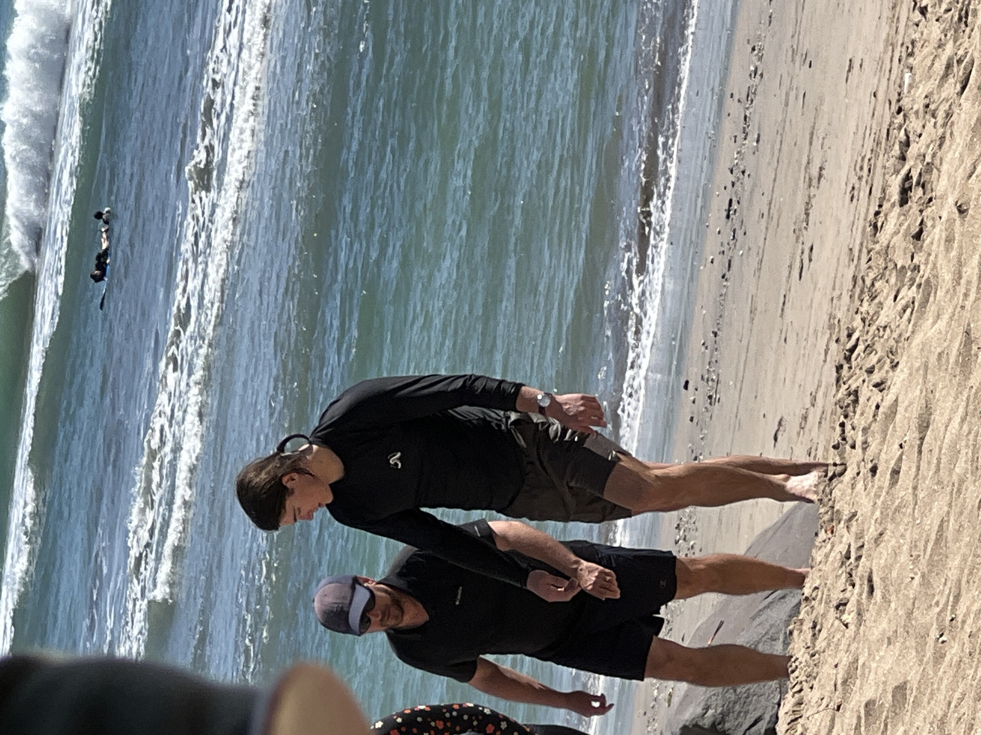 Two men are walking on a sandy beach. The man on the left is wearing a black polo shirt and black shorts, with a dark blue baseball cap and sunglasses. The man on the right is wearing a long-sleeved black shirt and dark shorts. He has dark hair pulled back into a ponytail and is wearing a watch on his left wrist. In the background, the ocean has waves, and two people are on surfboards in the water. The location is identified as Kuta, Indonesia.