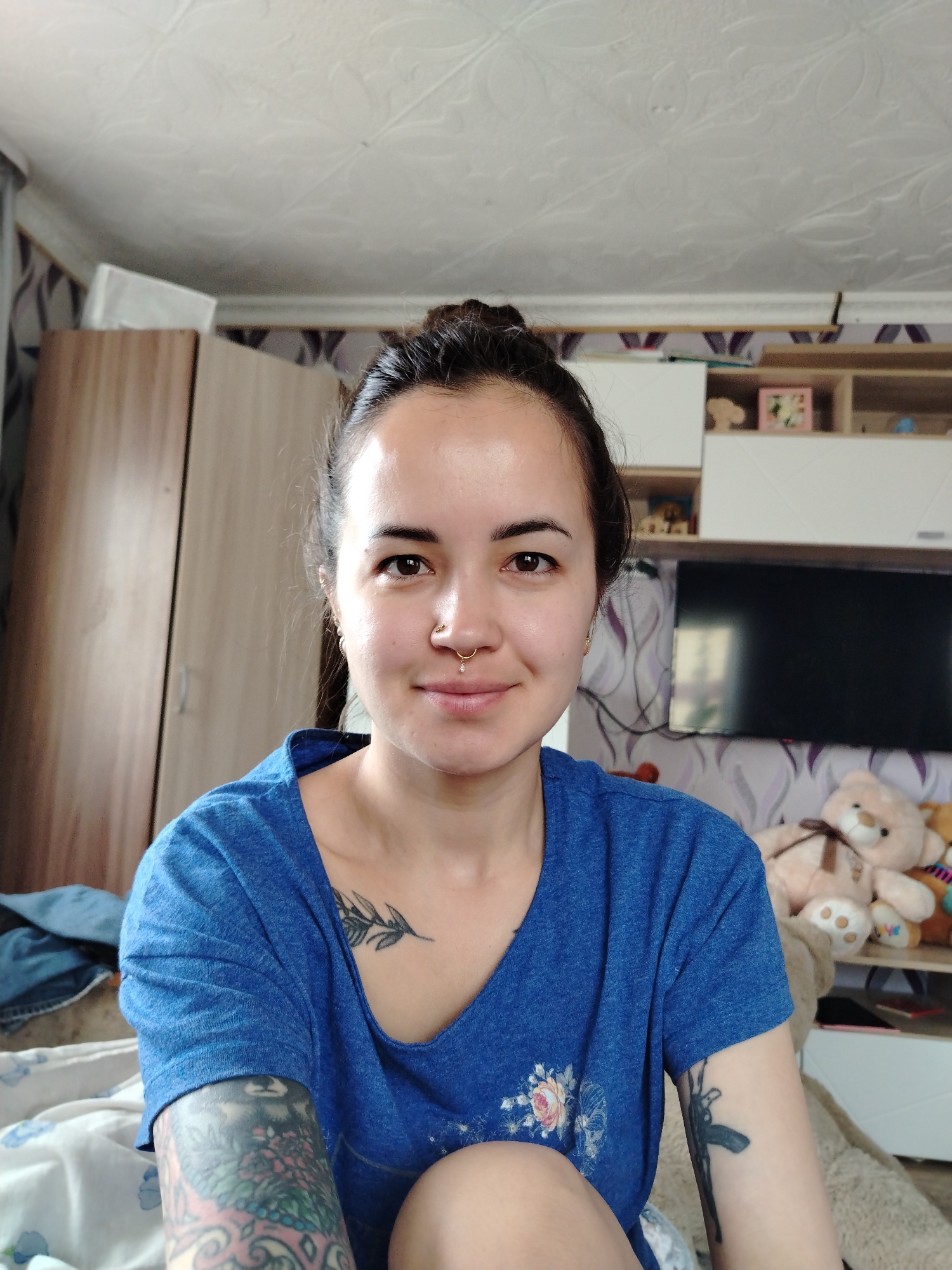 A woman is sitting indoors, presumably in a bedroom. She has dark hair pulled into a bun, brown eyes, and a nose ring. She is wearing a blue t-shirt and has tattoos on her arms and chest. The background includes a wardrobe, a TV, shelves with various items like toys, and patterned wallpaper. The location is most likely a residential building in Гожан, Russia.