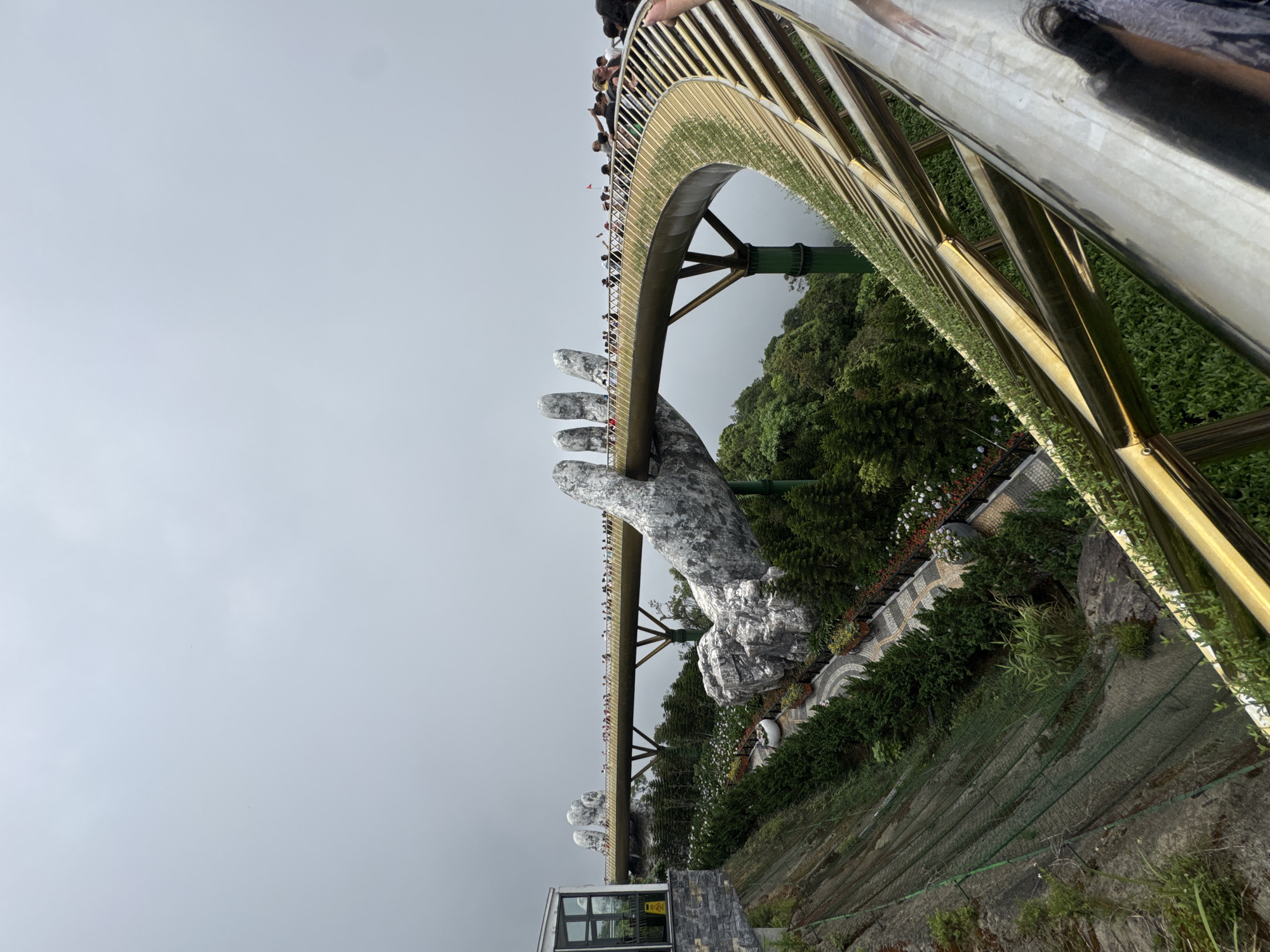 The image shows the Golden Bridge at the Bà Nà Hills resort near Đà Nẵng, Vietnam. The bridge is supported by two giant, stone-colored hands. People are walking along the bridge. The surrounding area is green with vegetation. The sky is overcast. Metal railings and support structures are visible in the foreground.