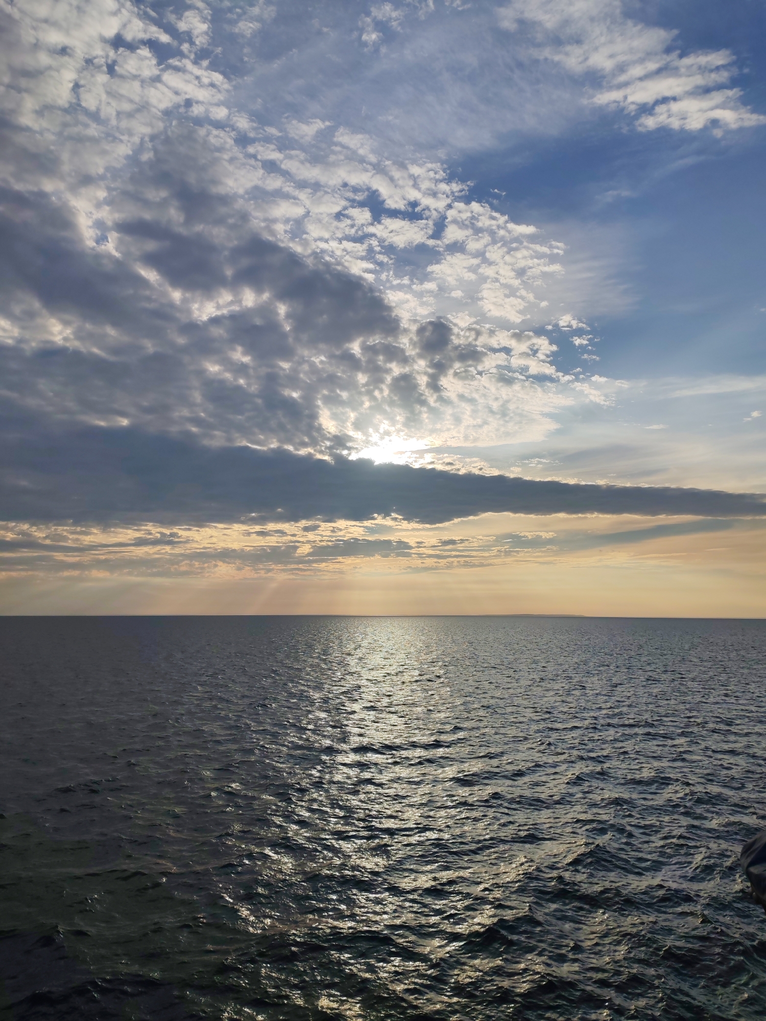 This is a landscape photograph taken at Ozernoe Ust'e, Russia, featuring a vast body of water under a dramatic sky. The horizon line is clearly defined, separating the dark, undulating surface of the water from the sky above. The water reflects the sunlight, creating a shimmering path across the surface. The sky is filled with a mix of clouds; some are dark and heavy, while others are wispy and white, allowing shafts of sunlight to break through. The overall impression is one of a serene, natural scene at dusk or dawn. There are no people or man-made structures visible in the photograph.