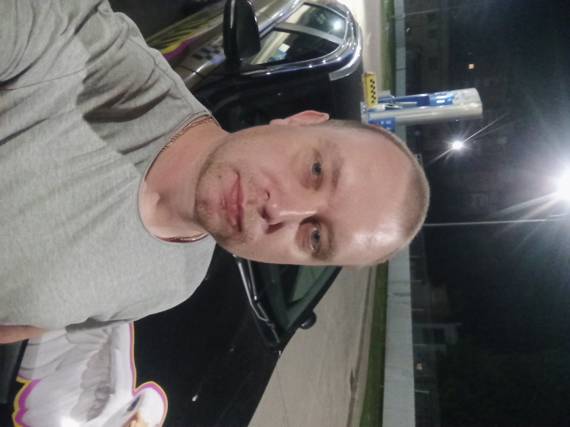 A man with a shaved head and fair skin is taking a selfie at night. He is wearing a grey t-shirt and a gold chain. Behind him, a black car with a taxi sign on the roof and a colorful decal of a bird on the hood is parked next to a gas station. The gas station features a tall, blue and white dispenser. The background includes a fence and some trees, suggesting an urban or suburban setting. The image appears to be taken in Mahilyow, Belarus, as indicated by the presence of a yellow taxi sign.