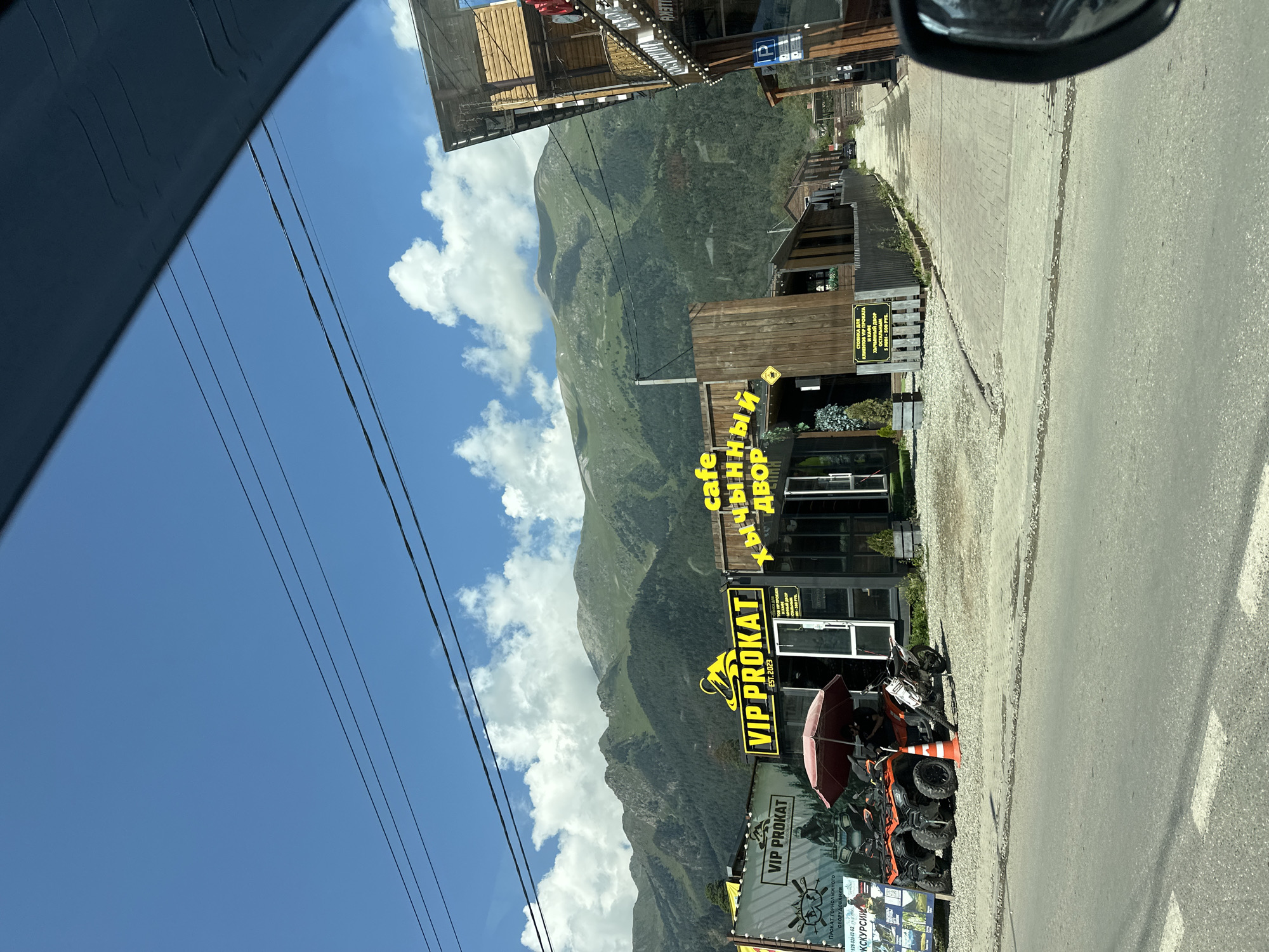 In the mountain town of Arkhyz, Russia, on a sunny day with a bright blue sky dotted with white clouds, a street scene unfolds. In the foreground, a gray asphalt road stretches towards a cluster of businesses. To the left, a building with large windows displays "VIP PROKAT," with ATVs and a motorcycle parked outside under a red umbrella, suggesting equipment rental services. Adjacent is another business labeled "cafe" with Cyrillic text in bright yellow neon lights, "Хычный Двор" which translates to "Khychinny Dvor" or "Khychin Courtyard," a local type of flatbread. The buildings feature a rustic wooden aesthetic, and a black fence lines the property. Further down the street, more wooden structures and a parked dark car are visible. Towering in the background are lush green mountains, their slopes covered in dense forest, reaching towards the sky. Power lines stretch across the frame. The scene captures the essence of a recreational mountain resort town.