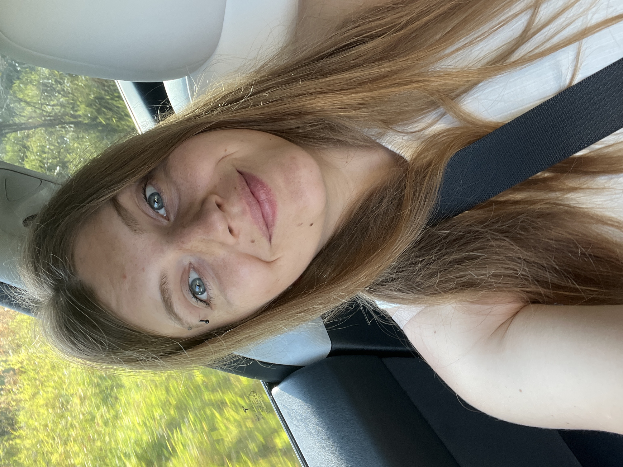 A young woman with long, auburn hair is taking a selfie inside a car, with a blurred green landscape visible through the window. She has blue eyes, freckles, and a small eyebrow piercing. She is wearing a light-colored top and a black seatbelt is visible across her chest. The interior of the car appears modern, with a white headrest visible. The image is taken from a low angle, focusing on the woman's face and upper body. The scene is outdoors, possibly on a highway given the blurred background indicating motion.