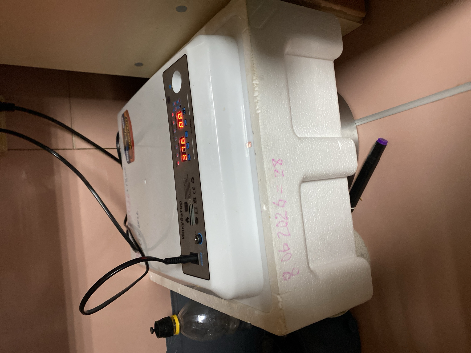The image shows a white incubator with a digital display showing "37.6" and "8.8". The incubator is sitting on a white styrofoam base. There is a clear plastic bottle with a black cap and yellow seal to the left of the incubator. Two black cords are plugged into the top of the incubator and extend upwards. Pink writing, which appears to be a date "8.06.2026" and the number "28", is written on the styrofoam base. A purple-tipped marker lies on the surface below the incubator. The background consists of a pinkish-brown tiled wall and a light brown cabinet. The overall setting appears to be an indoor, domestic environment, possibly in the city of Chirsha-Tartysh, Russia, given the text on the incubator which is in Cyrillic.