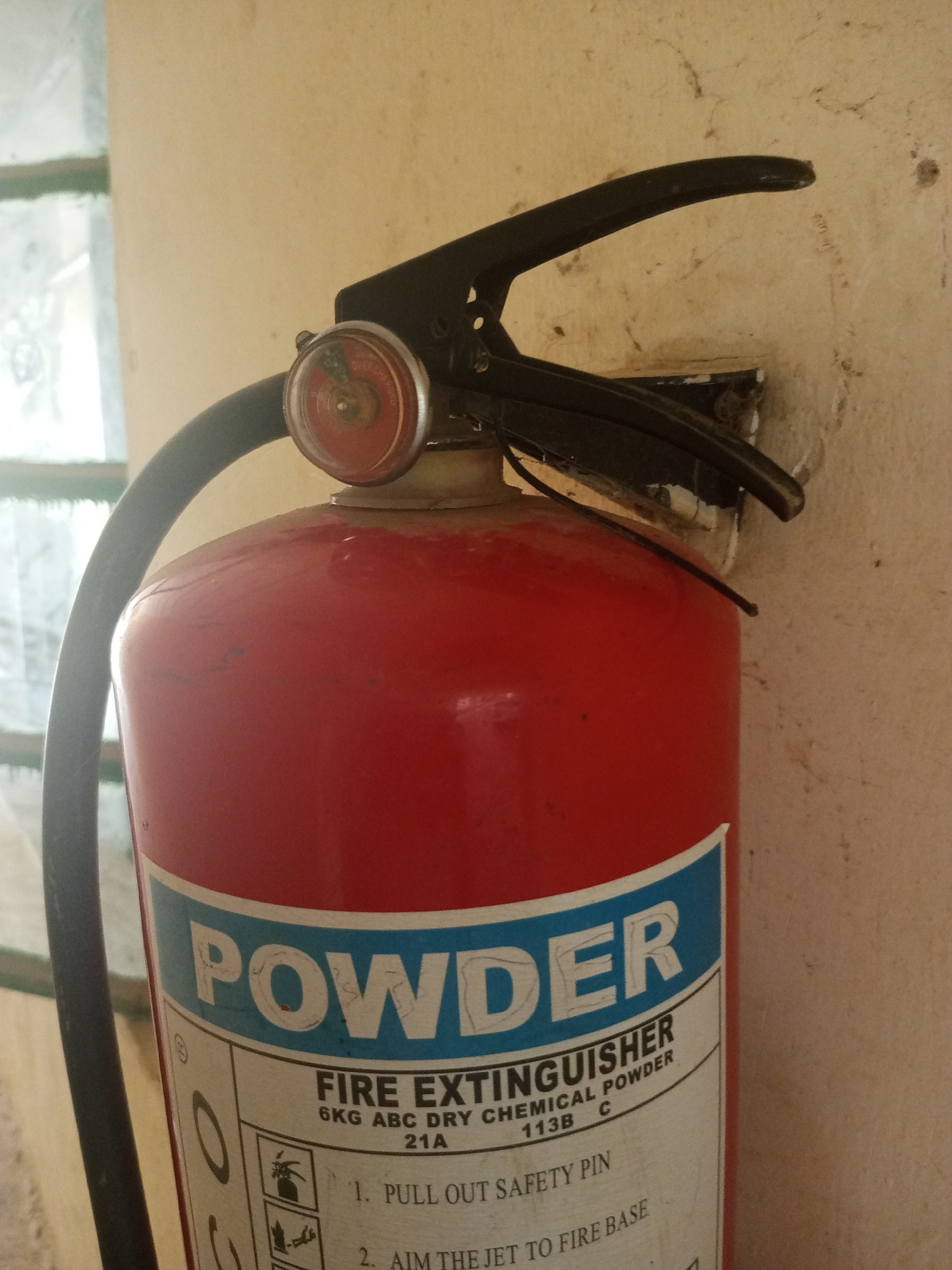 This media file is a close-up shot of a red fire extinguisher mounted on a textured, light-colored wall. The fire extinguisher is the primary subject, taking up most of the frame. Its label, predominantly white with blue lettering, is clearly visible. The word "POWDER" is written in large, bold, blue letters with white outlines across the top of the label. Below this, smaller text reads "FIRE EXTINGUISHER" and "6KG ABC DRY CHEMICAL POWDER". Further down, there are instructions with numbered steps. Step 1 reads "PULL OUT SAFETY PIN" and Step 2 reads "AIM THE JET TO FIRE BASE".
A pressure gauge with a red and green indicator is visible at the top of the extinguisher, just below the black handle. The handle itself is also black and appears to be made of plastic. To the left of the extinguisher, a portion of a window or glass partition is visible, with blurred green elements and light filtering through, suggesting it might be daytime. The wall behind the extinguisher shows signs of age and some dirt or smudges. The overall scene appears to be indoors, possibly in a hallway or a storage area. No people are visible in the image. The context provided is Jalingo, Nigeria.