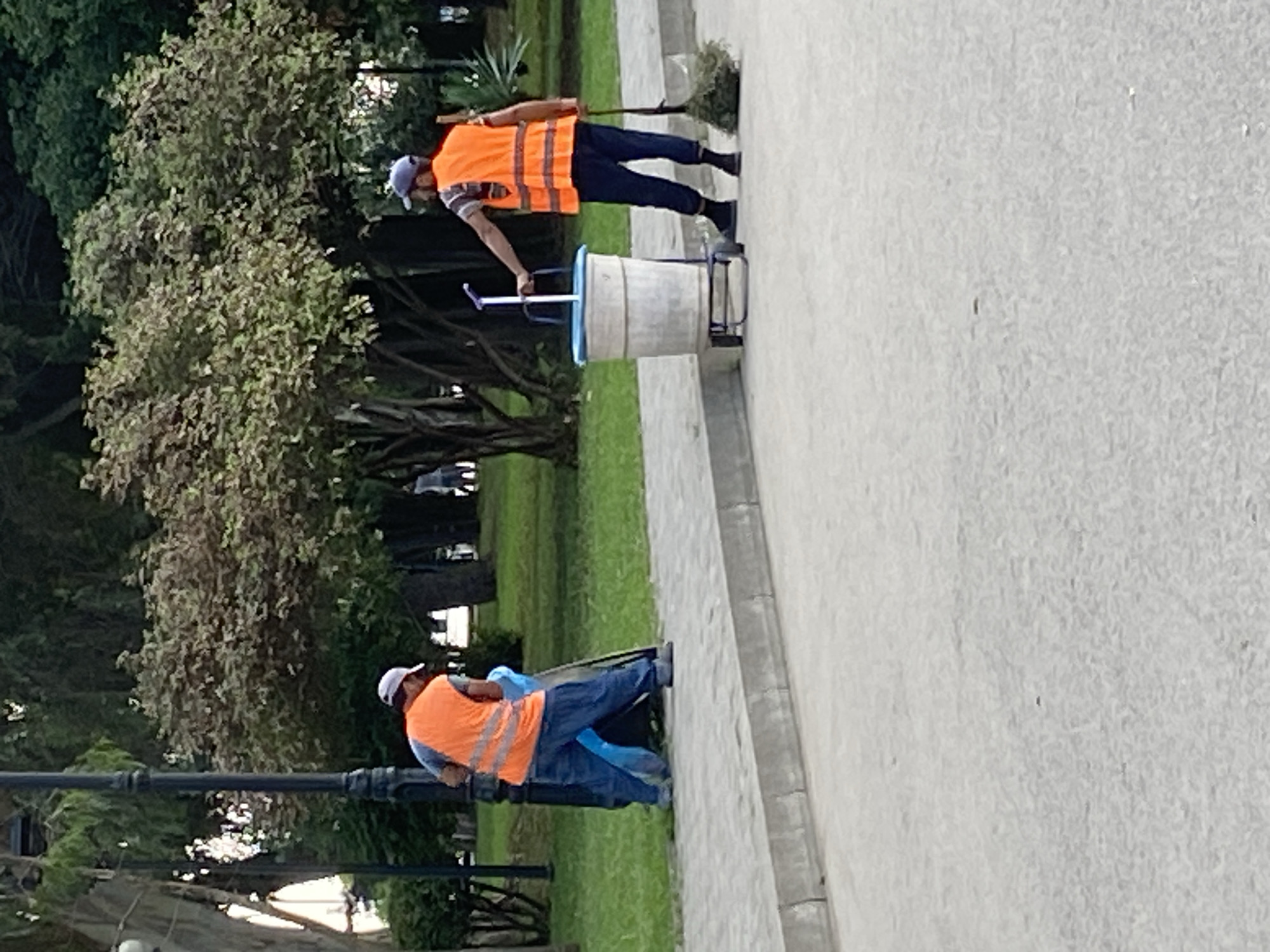 In a park in Sokhumi, Abkhazia, two sanitation workers are seen tending to the grounds. The worker on the left, wearing a white cap and an orange reflective vest over a grey shirt and blue jeans, is using a broom to sweep debris into a blue plastic bag. The worker on the right, wearing a blue cap and an orange reflective vest over a striped shirt and dark pants, is holding the handle of a wheeled garbage bin and stands next to a broom with a bundle of dried branches. The environment consists of a paved walkway, a grassy area, and trees. The scene appears to be illuminated by natural daylight.