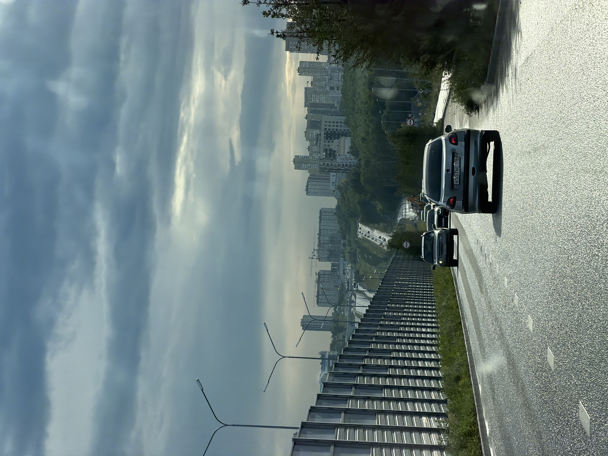 The image captures a view from a vehicle traveling on a highway, likely in an urban or suburban area of Kazan, Russia, given the architectural style of the buildings. The sky is overcast with dramatic, textured clouds, and shafts of sunlight pierce through, creating a dynamic atmosphere. On the left, a tall, grey sound barrier runs parallel to the road. Several streetlights with modern, angular designs are visible along the highway. In the distance, a dense collection of apartment buildings, some with unusual architectural features, dominates the skyline, suggesting a residential district. A lush green forest separates the highway from the city buildings. The road surface appears wet, indicating recent rain or damp conditions, and several cars are driving in the same direction. The primary subject in the foreground is a silver hatchback car, a Peugeot, with its rear facing the camera. The car's license plate reads "K557HC 116 RUS." The overall impression is one of transit through a developing urban landscape under a moody sky.