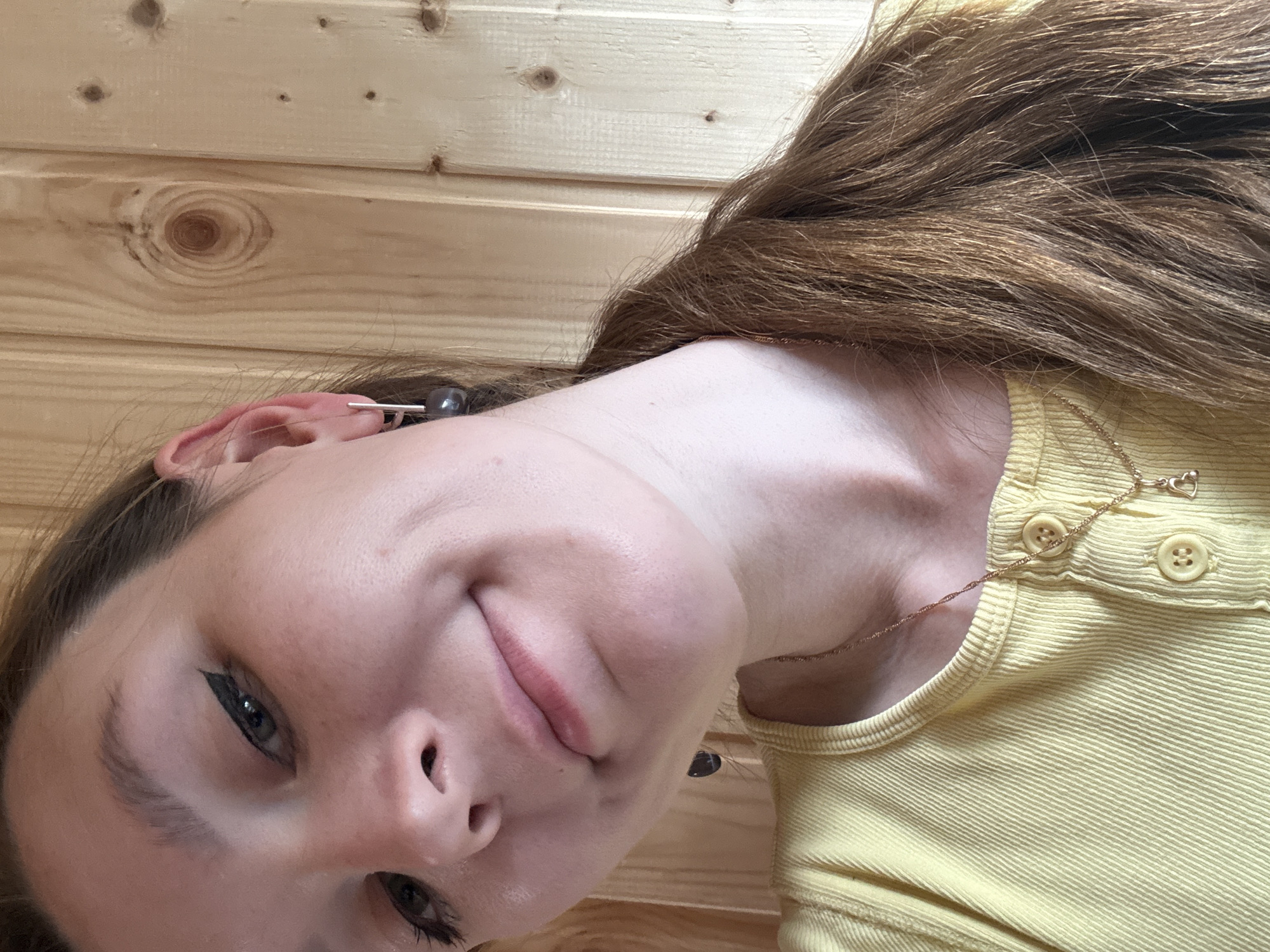 A young woman with long, brown hair pulled into a ponytail poses for a selfie in front of a wooden wall. She is wearing a yellow, ribbed t-shirt with two buttons and a delicate gold necklace with a small heart pendant. Her eye makeup is dramatic, with a winged eyeliner. The background appears to be an interior wall made of vertical wooden planks. There is no indication that this image was taken in the city of Italmas, Russia.