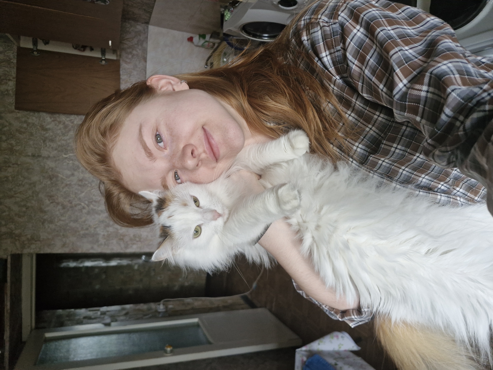 A young, fair-skinned woman with red hair, likely in her late teens or early twenties, is holding a large, fluffy white cat with patches of brown and black on its head. The woman is wearing a brown and gray plaid shirt. The background appears to be a domestic interior, possibly a kitchen, with cabinets and a window visible. The exact location in Yekaterinburg, Russia, cannot be determined from the provided image.
