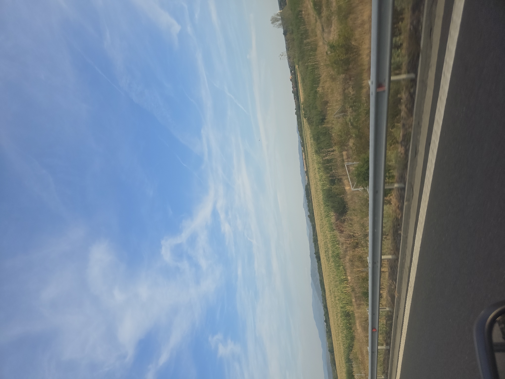 The image depicts a landscape view from a moving vehicle, likely on a highway or a road in the vicinity of Susani, Romania. The foreground features a dark asphalt road with a white stripe and a metallic guardrail. Beyond the guardrail, there is a field of crops, appearing to be corn, with some dry grass and vegetation. In the background, rolling hills or mountains are visible under a bright blue sky with scattered wispy white clouds and contrails. The overall scene suggests a rural or semi-rural setting during daylight hours, possibly in late summer or early autumn given the appearance of the crops. There are no people or discernible actions depicted in the image.