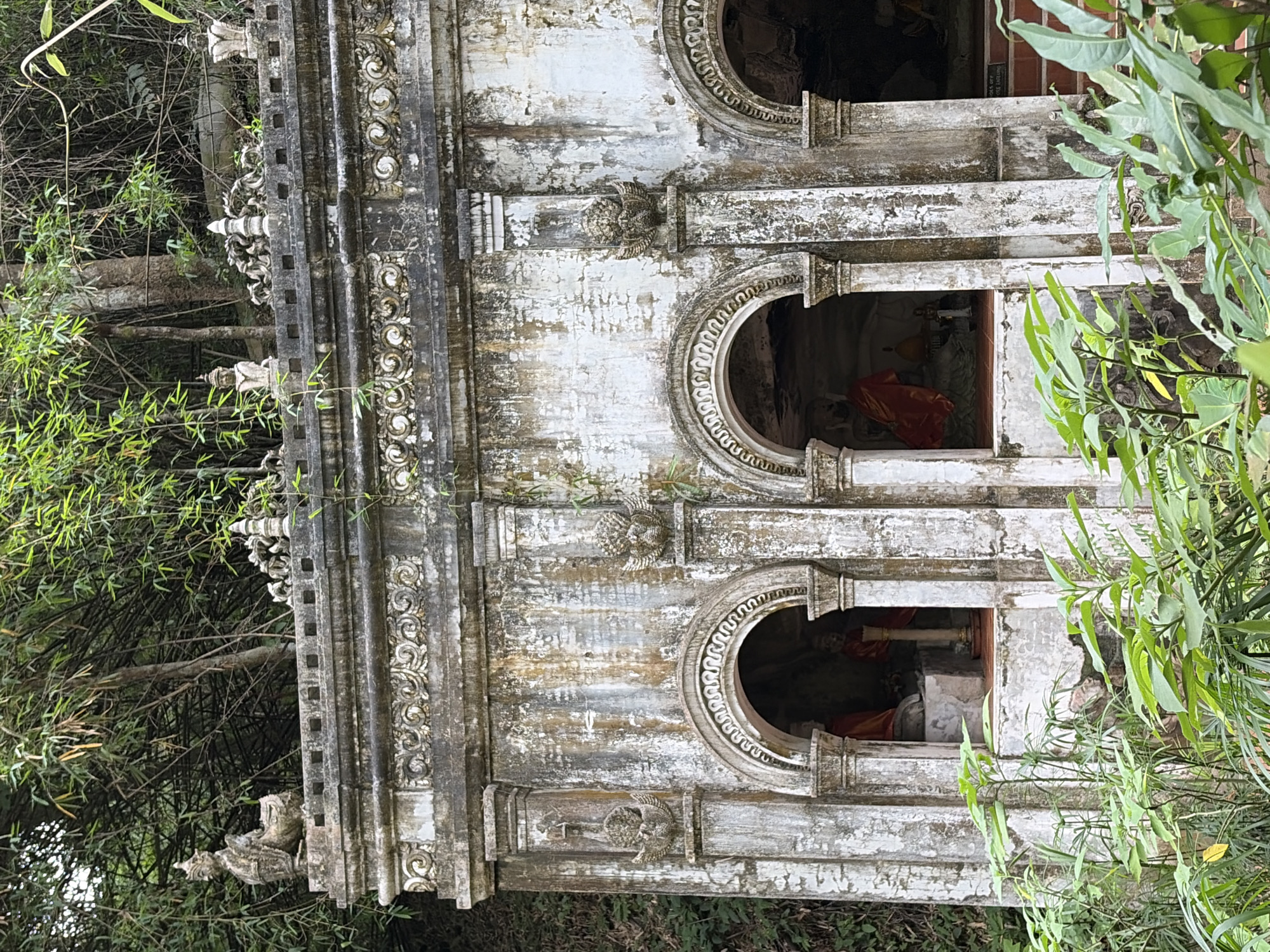 The image shows the exterior of an old, weathered building with ornate architectural details. The building is made of concrete or stucco and is stained with age and moisture. It features multiple arched openings, each appearing to house a statue or religious effigy. The statues are partially obscured and appear to be adorned with robes. The building is decorated with carved moldings, decorative elements on the pillars, and small statues on the roofline. Surrounding the structure are lush green foliage and trees, suggesting a natural or overgrown setting. The overall impression is one of antiquity and a spiritual or historical site, possibly within a temple complex. Based on external knowledge, this structure is located within the grounds of Wat Phra That Doi Suthep, a prominent temple in Suthep Town Municipality, Thailand, likely a shrine or annex within the larger temple complex. There are no discernible people in the scene.