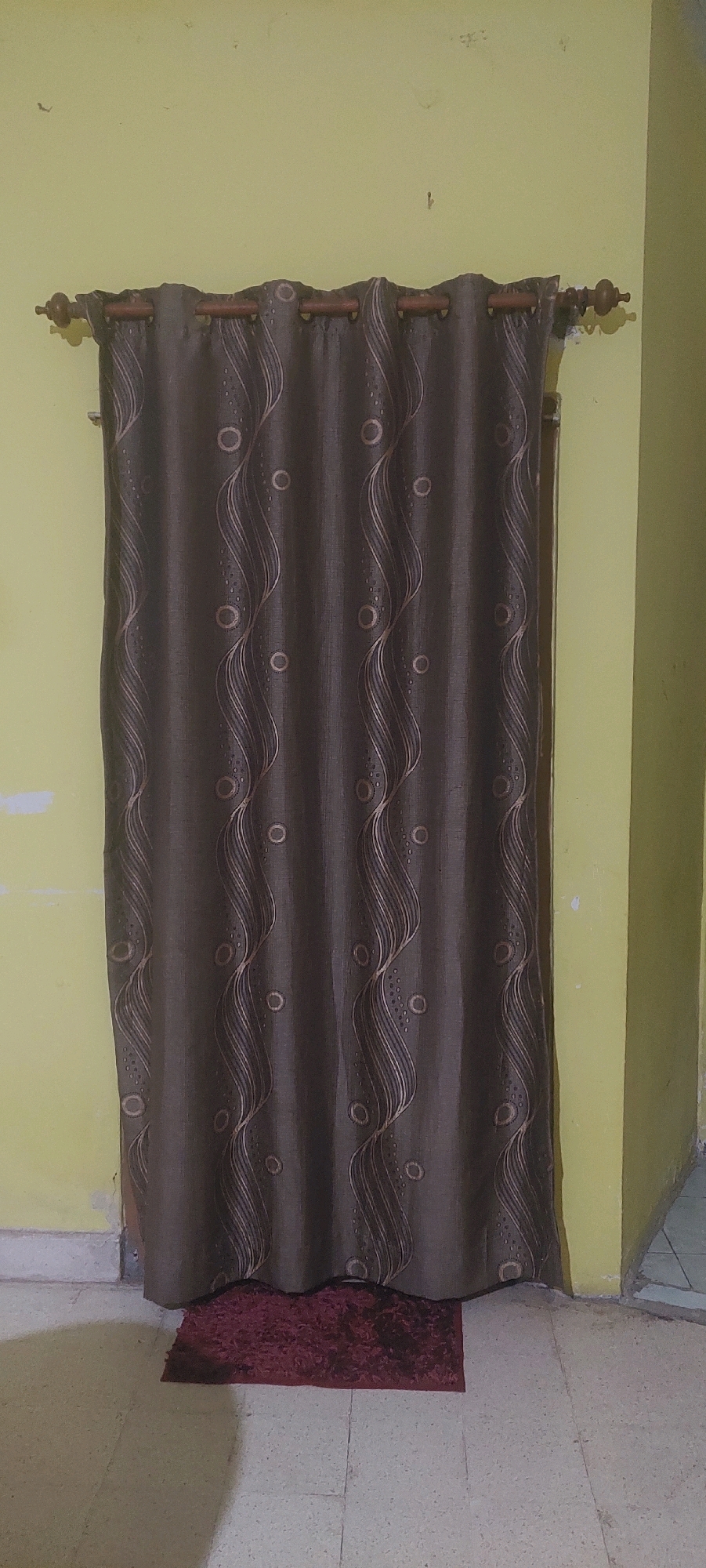 A dark brown curtain with a wavy pattern of lighter brown lines and circles hangs from a wooden curtain rod against a yellow wall. The curtain covers most of a doorway. A small, dark red rug is placed on the tiled floor directly in front of the curtain. The floor is made of large, square tiles. The wall appears to have some scuff marks and discoloration. The scene is indoors and appears to be a simple room. No people are visible in the image.
