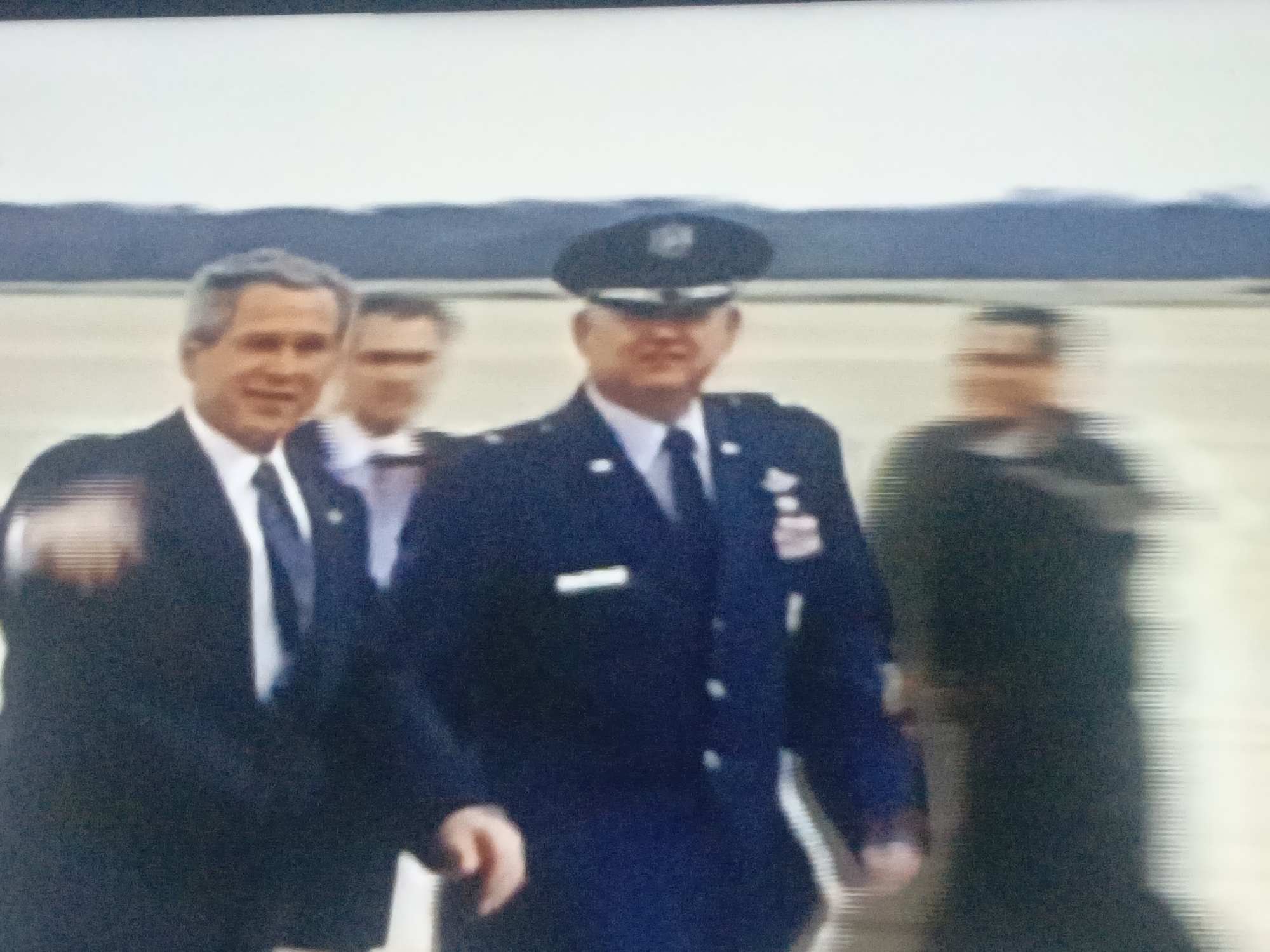 The image shows George W. Bush and several men walking outside on a paved surface, possibly an airfield. Bush is on the left, wearing a dark suit and tie. Next to him is another man in a suit. To their right, a man in a military uniform with a cap and various insignia is visible. Another blurred figure is to the right. The background shows a hazy, pale sky over land or distant hills. No elements suggest the location is Juan Griego, Venezuela.