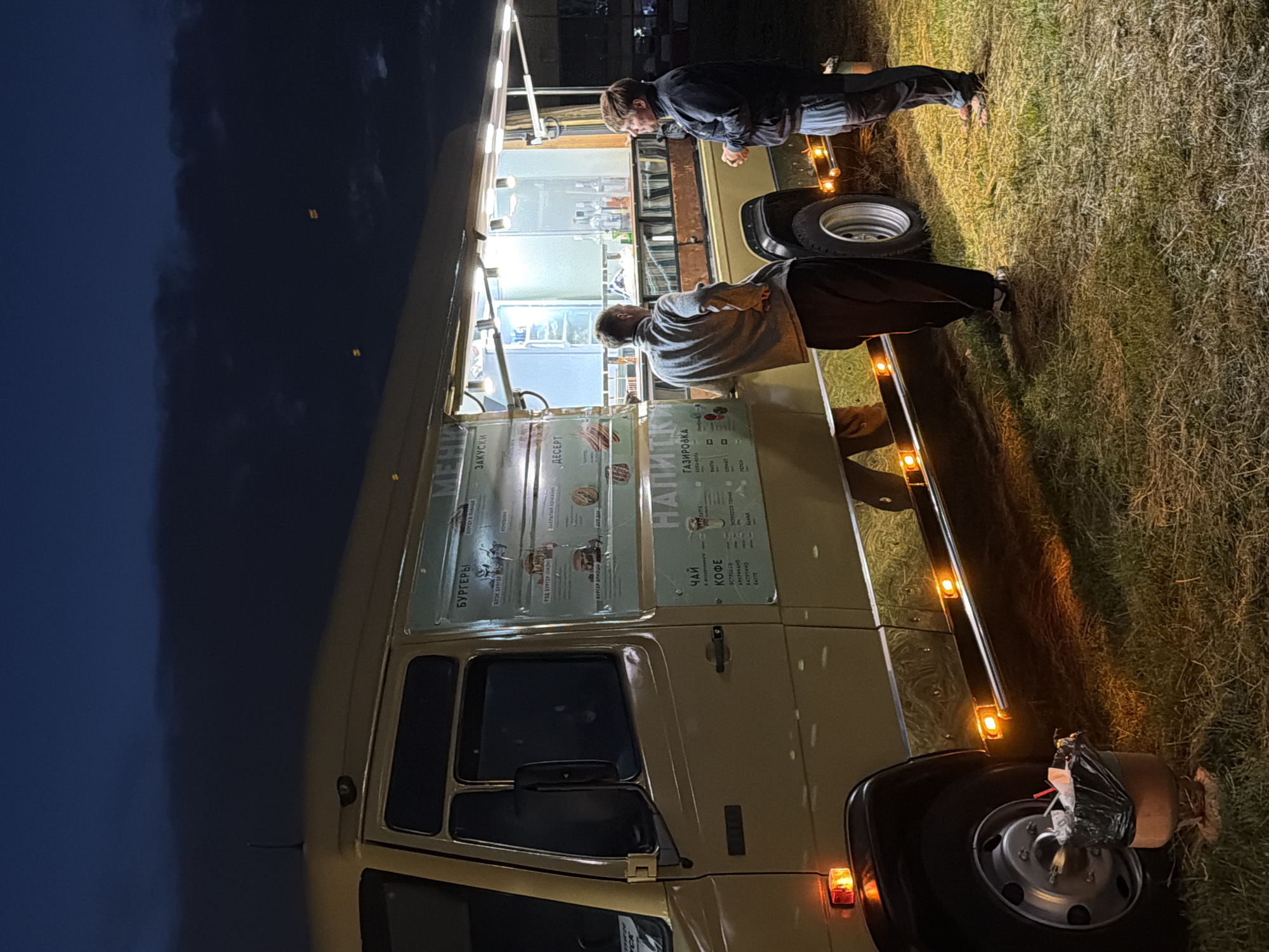 At night, a beige food truck is parked on a grassy area. Two men stand in front of the truck, one facing the truck and the other facing away from it. The food truck has a menu displayed on its side, with sections for "BURGERS," "SNACKS," "DESSERT," and "DRINKS." The menu lists items such as "TEA," "COFFEE," and "SODA." The interior of the truck is illuminated by bright lights. In the foreground, a trash can is visible next to the truck's wheel. The sky is dark, with some clouds visible.