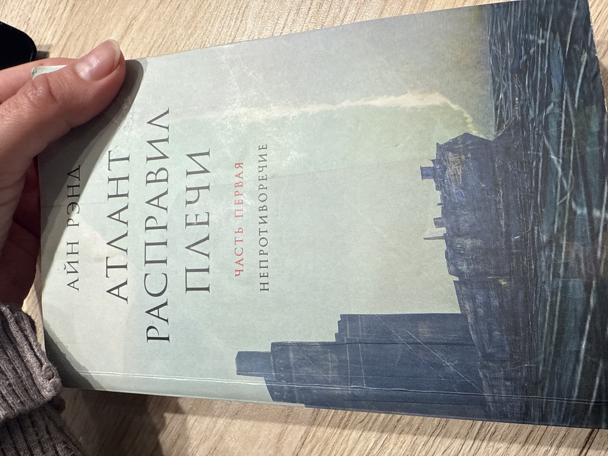 A person's hand holds a book, which is displayed against a light-colored wooden surface, likely a table or floor, suggesting an indoor setting. The hand, visible on the upper left, has light skin and well-maintained fingernails. The book's cover features a muted, desaturated color palette dominated by shades of grey, light green, and dark blue. The artwork depicts a stylized, somewhat abstract urban or industrial landscape, with dark, angular structures resembling buildings or silos silhouetted against a lighter sky. On the far right, a pattern of dark vertical lines suggests rain, reflections, or architectural details.
The text on the book cover is in Russian. Vertically aligned on the left side, the author's name reads АЙН РЭНД (Ayn Rand). The main title, also vertically oriented and prominent, is АТЛАНТ РАСПРАВИЛ ПЛЕЧИ (Atlas Shrugged). In the middle-right section, red vertical text indicates ЧАСТЬ ПЕРВАЯ (Part One), with a subtitle below it, НЕПРОТИВОРЕЧИЕ (Non-Contradiction). The image captures the simple act of holding or presenting the book. No specific time of day or emotional context is discernible from the scene.