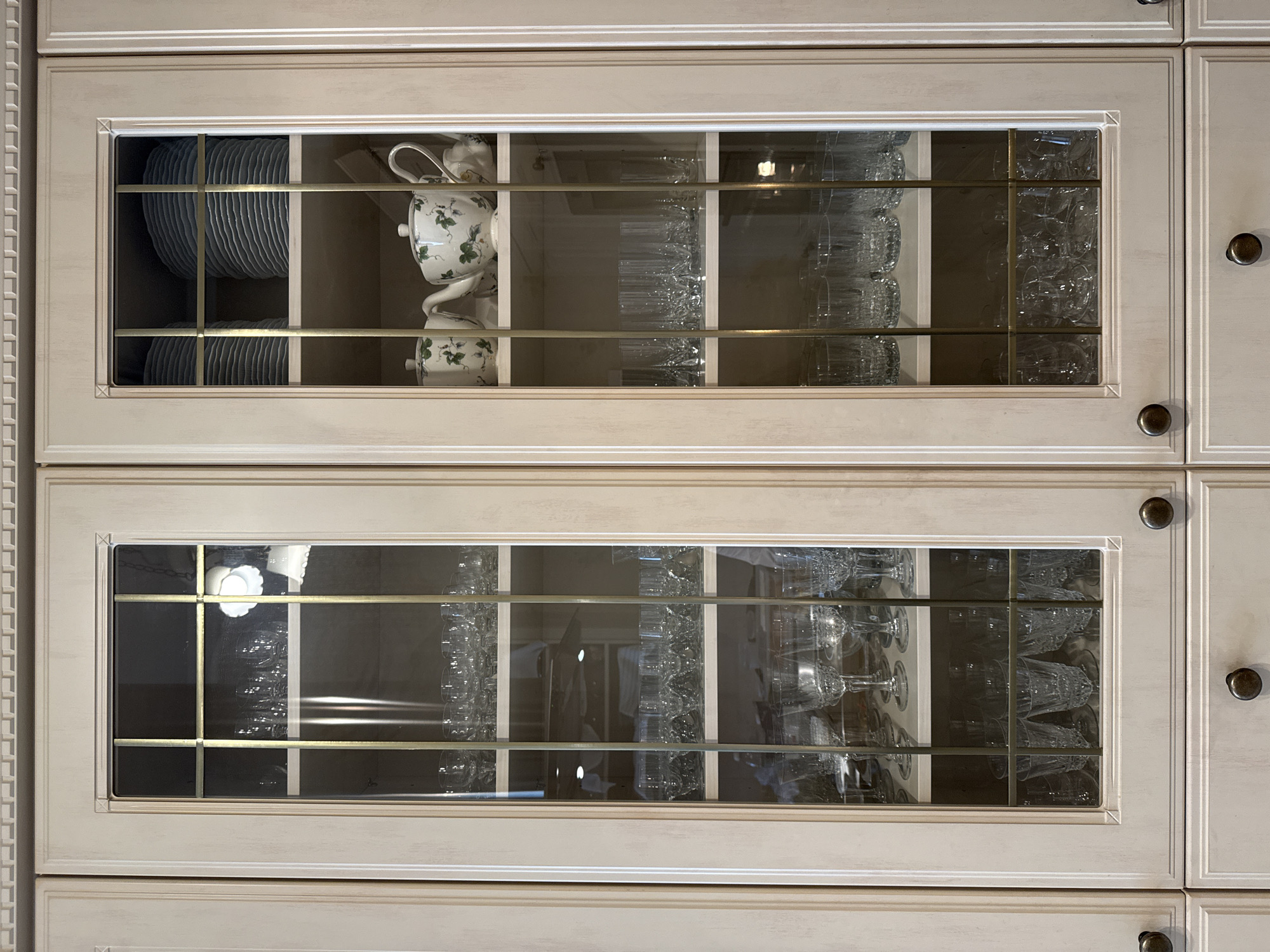 A close-up shot presents a section of light-colored kitchen cabinets with glass-paneled doors. The upper cabinet displays plates and a floral-patterned teapot with matching cups, as well as rows of glasses. The lower cabinet is filled with more glassware of varying shapes. The overall impression is of organized kitchenware displayed behind glass doors, set within the context of a domestic interior. There is no evidence in the photograph that indicates the location in Saint Petersburg, Russia.
