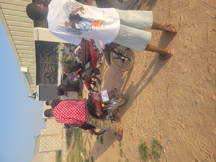 This image depicts an outdoor scene in Ibo, Nigeria, on a dry, unpaved dirt surface. Four male subjects and three motorcycles are visible.
In the foreground, a male subject stands facing away from the camera, positioned slightly right of center. He wears a white t-shirt with a graphic design of a winged figure on the back, light blue denim shorts, and brown flip-flops. His arms are not clearly visible.
To the left, a second male subject is seated on a red motorcycle, also facing away from the camera. He wears a red and white patterned long-sleeved shirt and camouflage-patterned trousers, with brown sandals on his feet. The motorcycle has a white license plate with red lettering, partially visible as "P-MPRE."
Behind these two individuals, near a light-colored single-story building with a corrugated metal roof and horizontal dark grey panels, two additional male subjects are present. One is partially obscured by the motorcycle; the other is bent over, facing the building, seemingly interacting with a fixture or plant located at its base. Both wear dark clothing.
Two other red motorcycles are partially visible: one to the right of the seated subject, and another mostly obscured by the standing subject on the right.
The ground is predominantly reddish-brown dirt or sand, showing numerous shoe prints and tire tracks, with sparse patches of green vegetation. Long shadows are cast on the ground, indicating a low sun angle. In the background left, an unfinished wall constructed from light-colored concrete blocks is visible. The sky is clear and light blue.