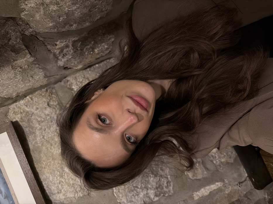 The image captures a female subject indoors, within the city of Morinj, Montenegro. The woman has long, wavy dark brown hair, light blue-green eyes, and is wearing a brown or taupe blazer over a dark garment. A small gold stud earring is visible in her right ear. She is looking directly towards the camera with a subtle, closed-mouth smile. The background features a rustic stone wall composed of irregularly shaped grey and brown stones with visible mortar. In the upper left quadrant, a framed picture or artwork with a dark frame and a light mat is partially visible against the stone. The lighting in the scene is subdued.
