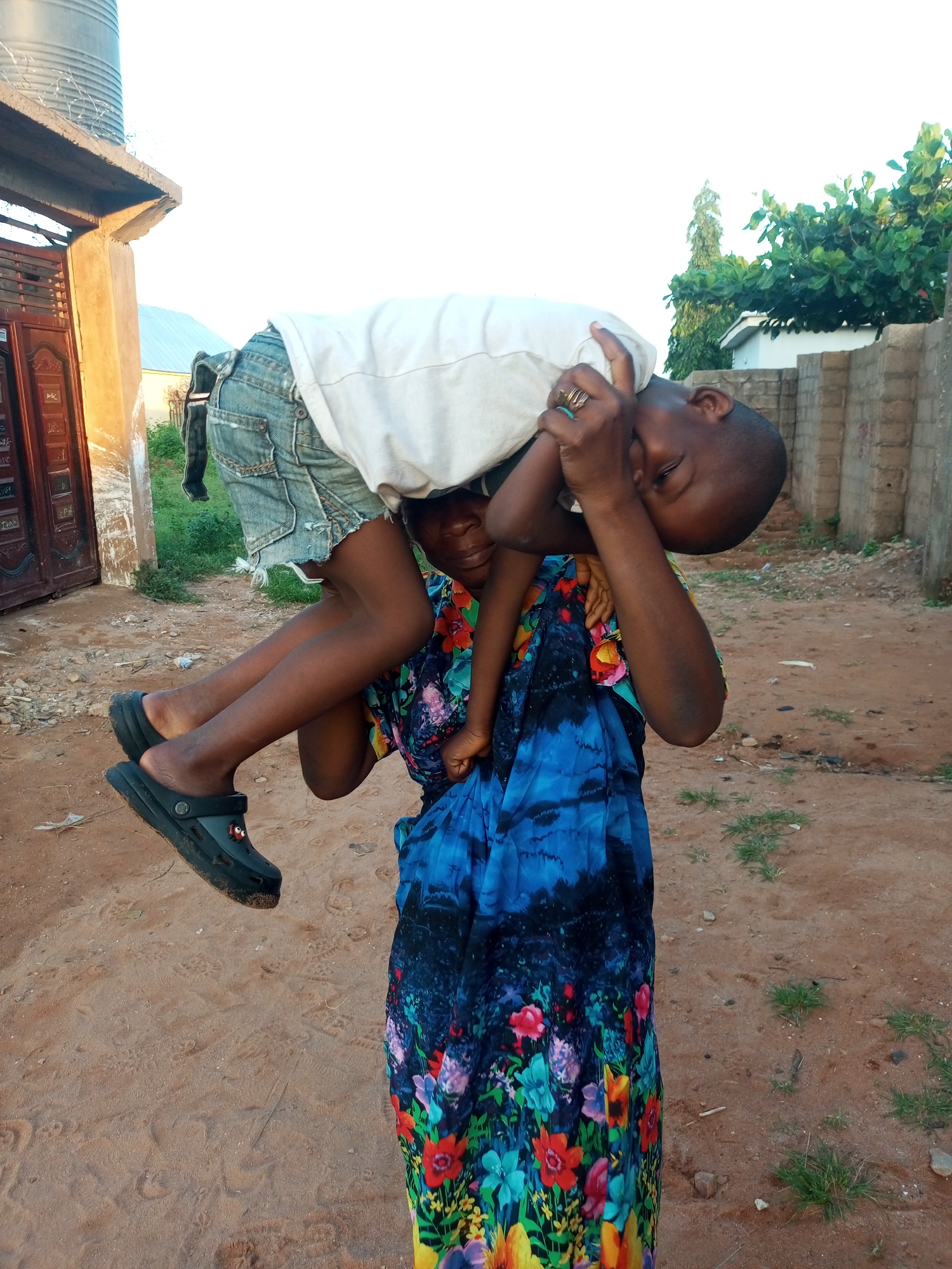 In Tempure, Nigeria, a person carries a child on their shoulders. The child, wearing a white t-shirt and denim shorts, is held upside down with their head tilted back. The person carrying the child is wearing a vibrant blue and floral print dress. The ground is a mix of sand and dirt, with some sparse green vegetation. In the background, a brick wall and some trees are visible. A large water tank and part of a building are also present on the left side of the image.