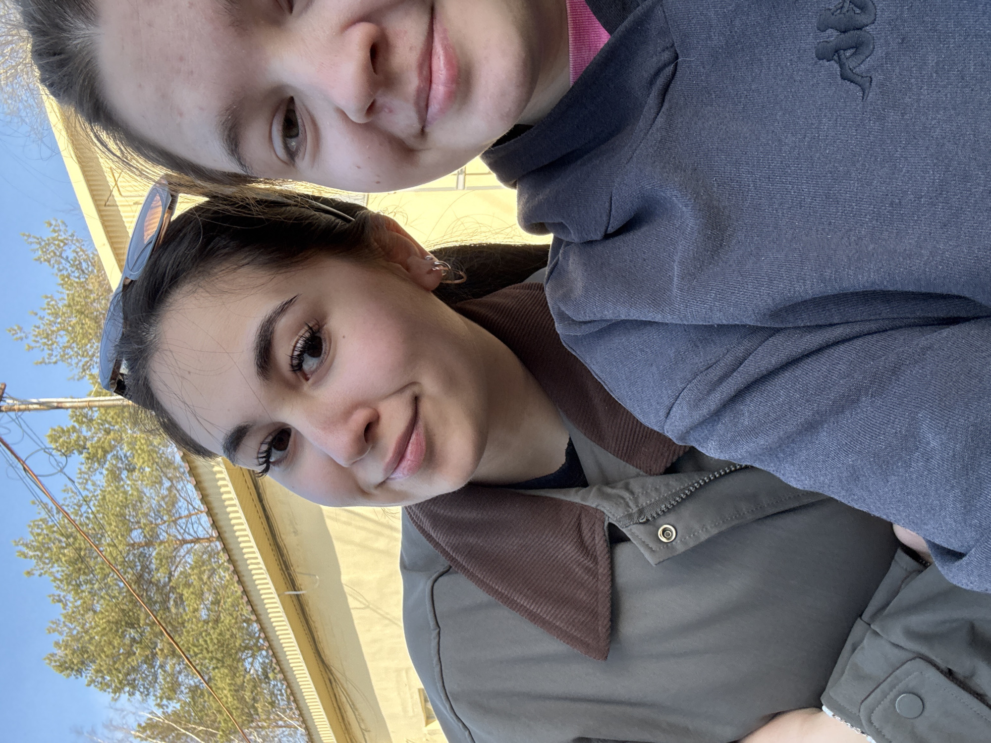 Two young women are posing closely for a selfie outdoors on a sunny day. The woman in the foreground is wearing an olive-green jacket with a brown corduroy collar. Sunglasses are perched on her head. The other woman is wearing a gray hoodie with a snake logo on the chest. The background shows a yellow building, trees, and a blue sky with overhead electrical wires. It is day outside. The location is consistent with the city of Chaikovsky, Russia.