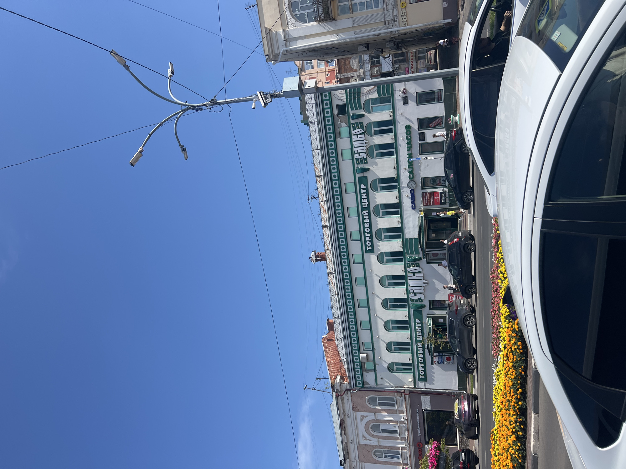 A sunny day in Ulyanovsk, Russia, shows a street scene with a multi-story building dominating the frame. The building, with its white facade and green trim, appears to be a shopping center, as indicated by the "ТОРГОВЫЙ ЦЕНТР" (Shopping Center) signage in large green letters. Below this, a sign for "CASIO" and "САЛОН ЧАСОВ" (Watch Salon) is visible. Several cars are parked or driving on the street in front of the building. A man is visible inside the shopping center, and other pedestrians are on the sidewalk. A bright flower bed with yellow and orange flowers lines the street. The sky is clear blue with a few wispy clouds. A street lamp with multiple lights and a security camera is positioned on the right side of the image.